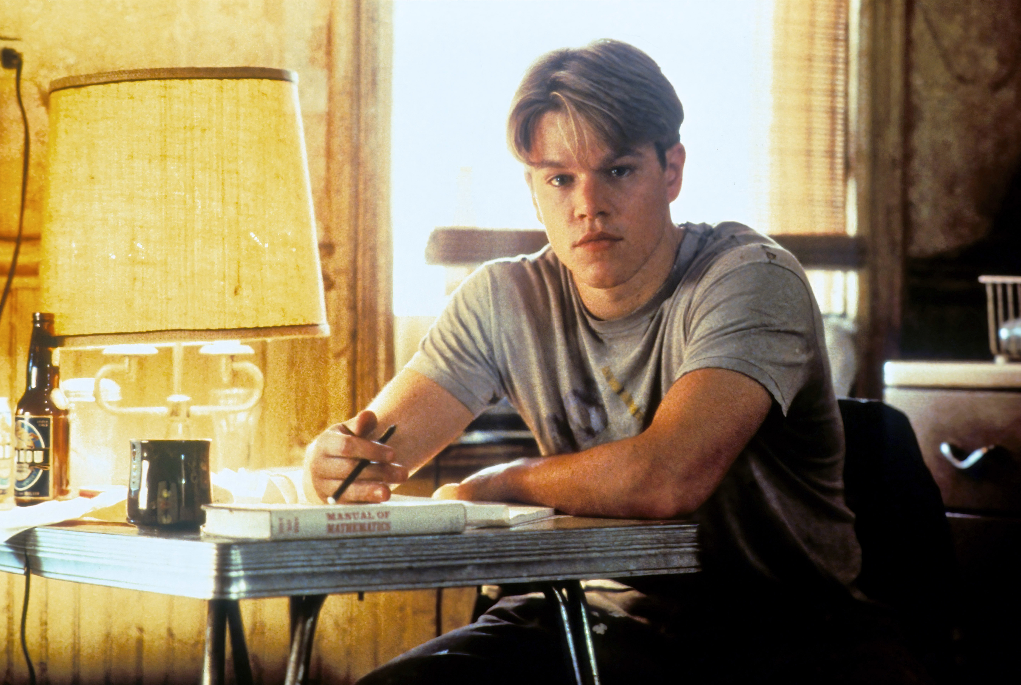 Matt Damon, in a casual t-shirt, sits at a table with a study book titled &quot;Manual of Mathematics&quot;, a pen in hand, looking towards the camera