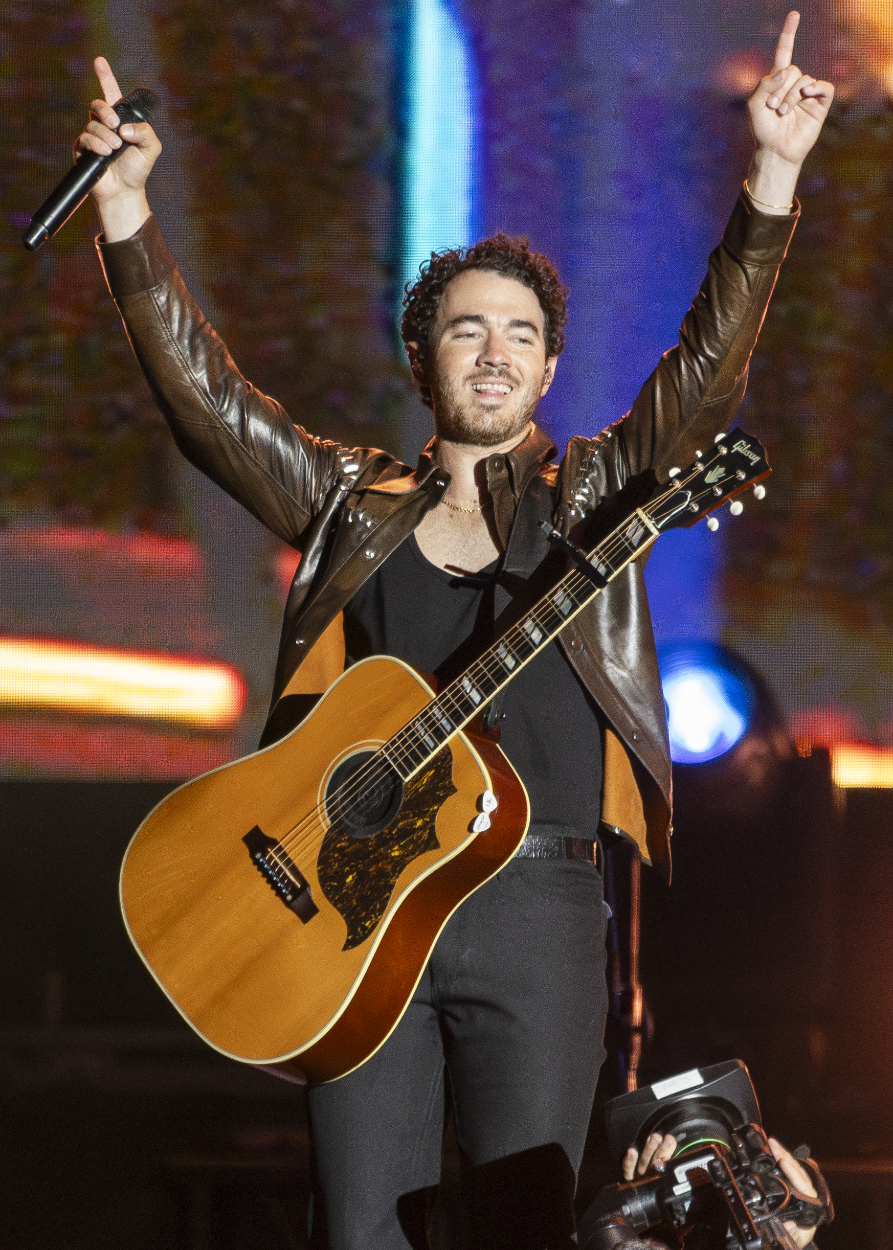 Kevin Jonas performs on stage, wearing a black shirt, black pants, and a brown leather jacket, holding a guitar and raising his hands