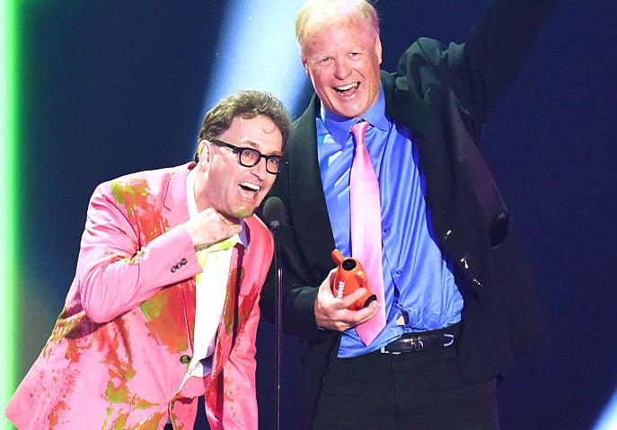 Stephen Colbert and Ed Begley Jr. on stage, with Ed wearing a suit and pink tie and Stephen in a colorful suit, sharing a moment at a celebrity event