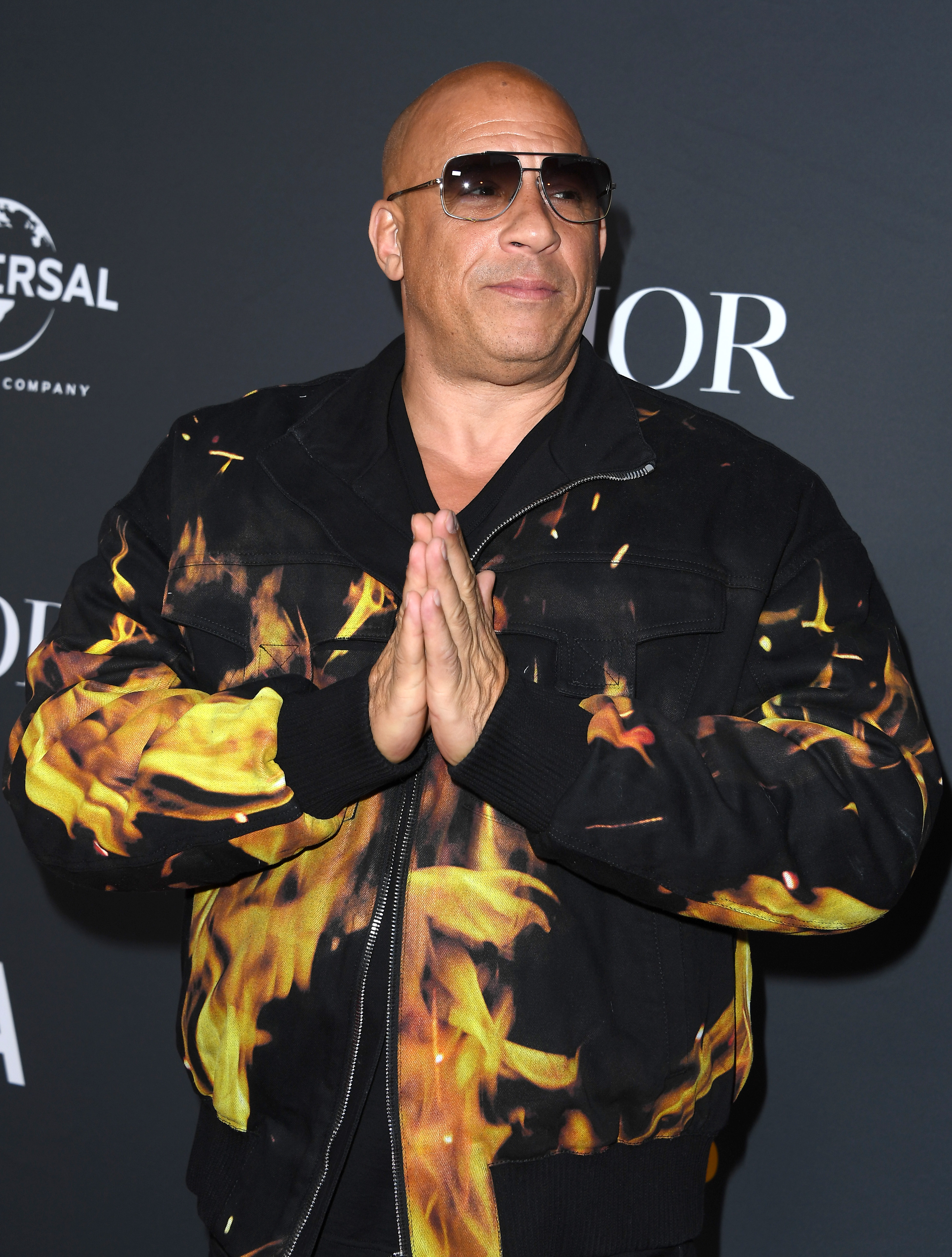 Vin Diesel on a red carpet wearing sunglasses and a jacket with a flame design, hands clasped