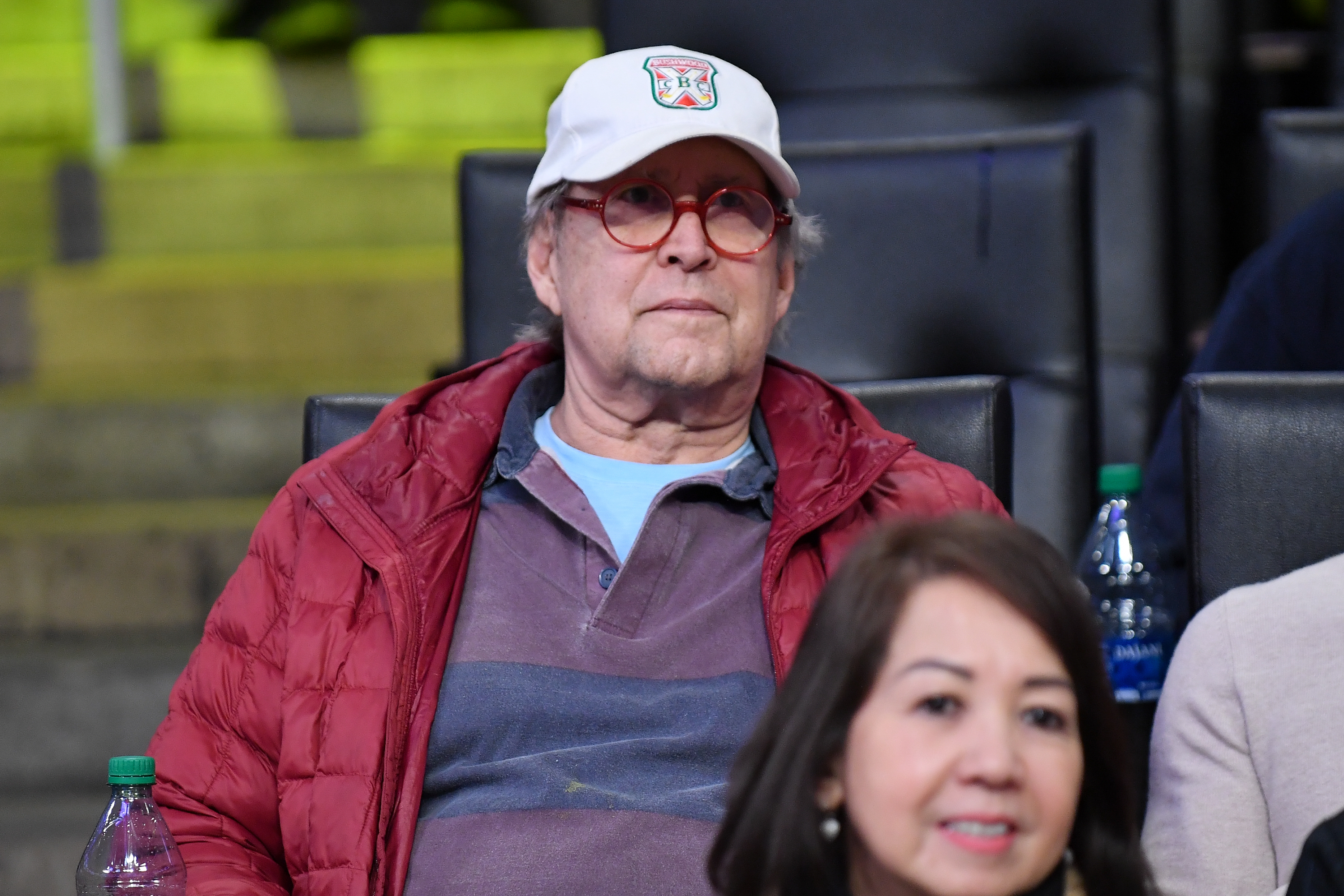 Chevy Chase at an event, wearing a white cap and glasses, with a red jacket over a blue shirt