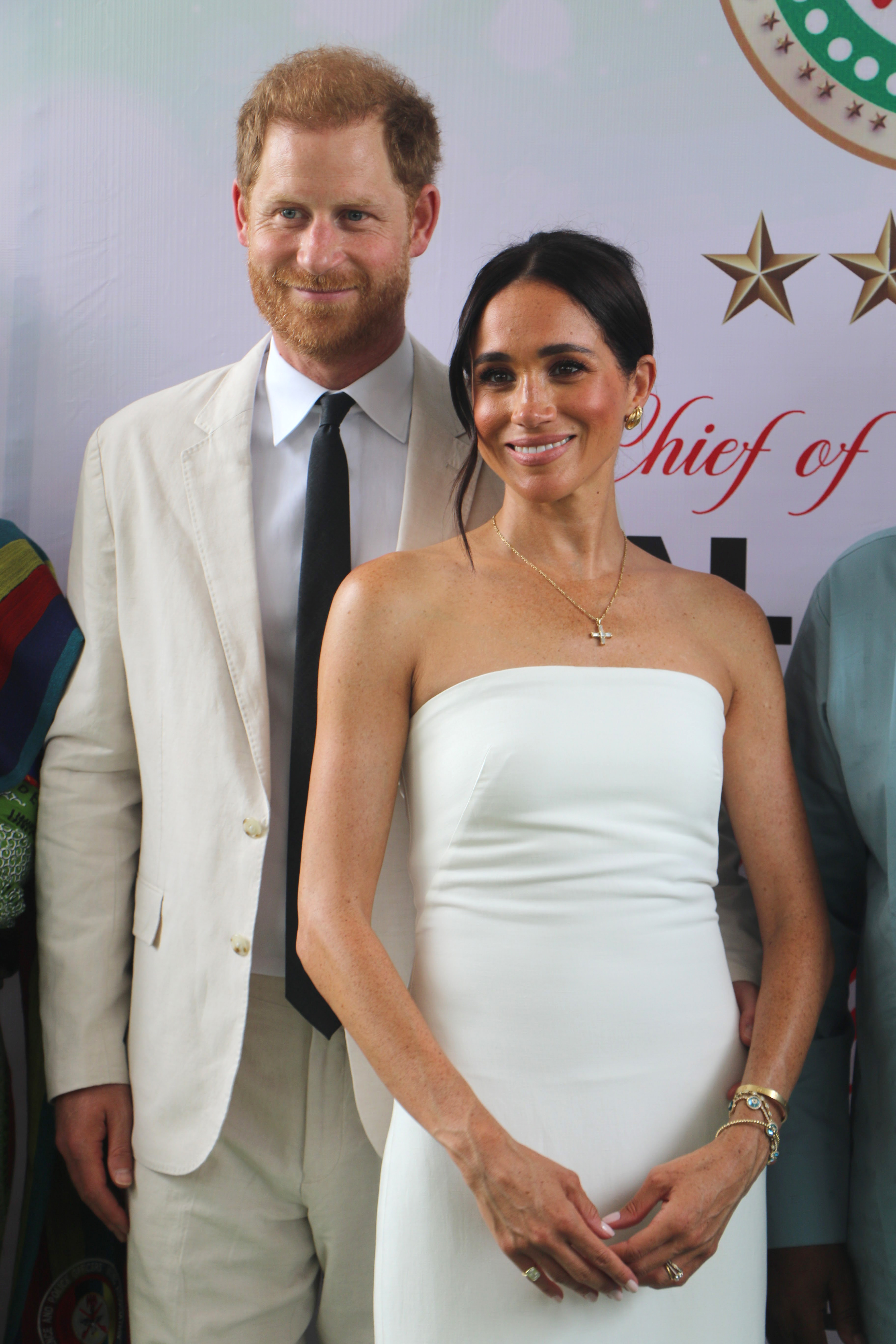 Prince Harry in a beige suit and Meghan Markle in a strapless white dress posing together at a public event