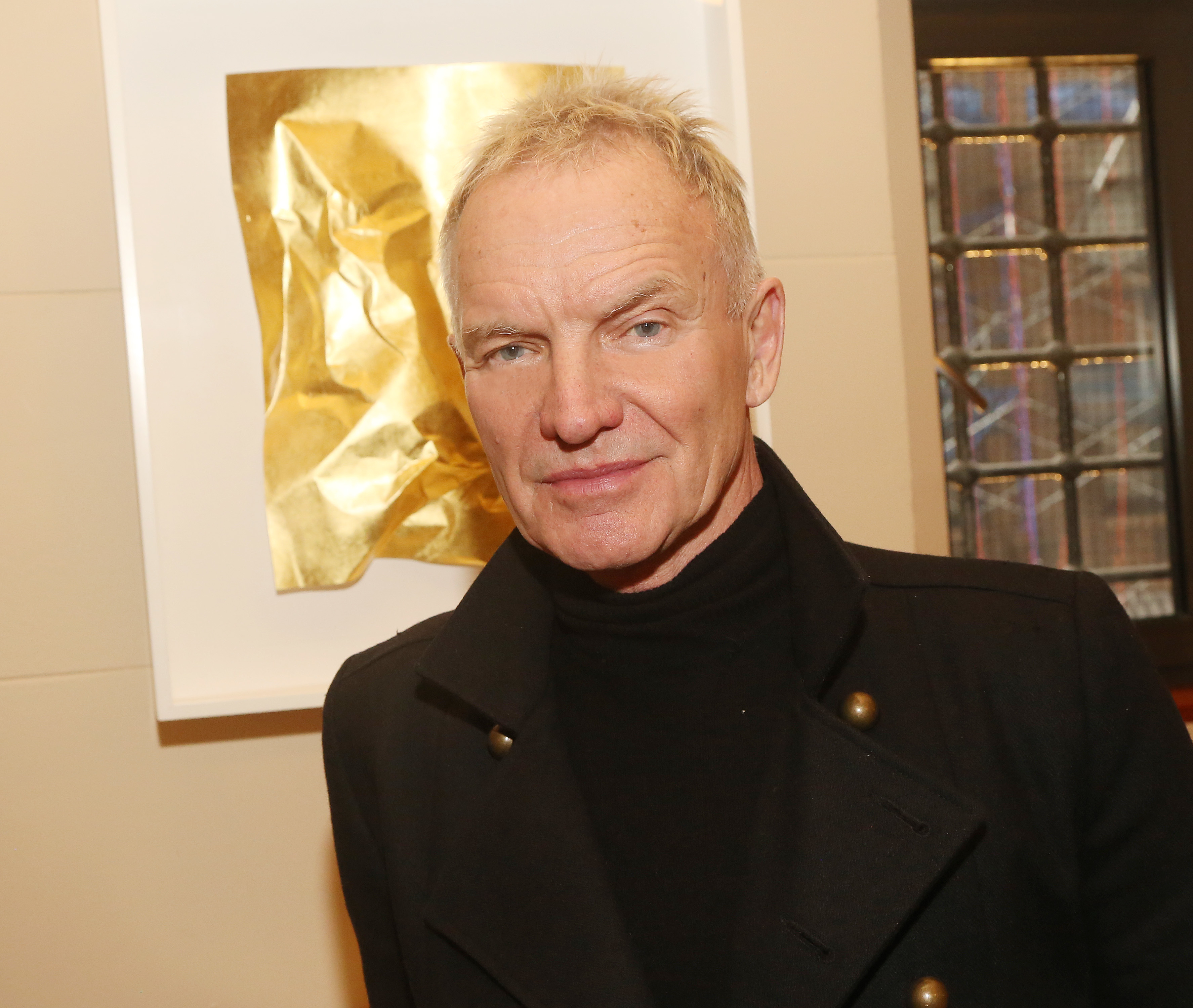 Sting, wearing a black turtleneck and black coat, stands in front of framed artwork