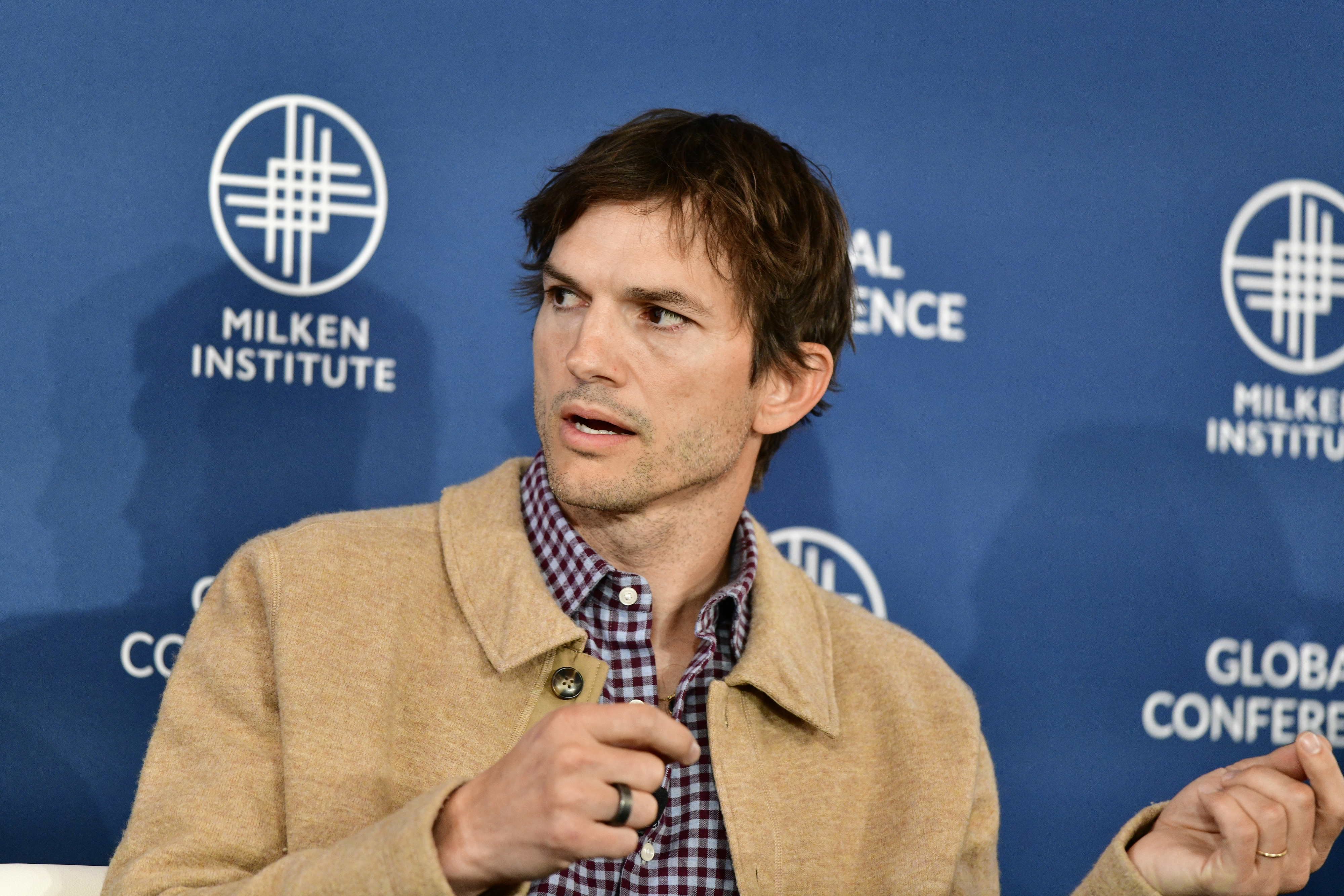 Ashton Kutcher speaks at the Milken Institute Global Conference, wearing a beige jacket over a checkered shirt