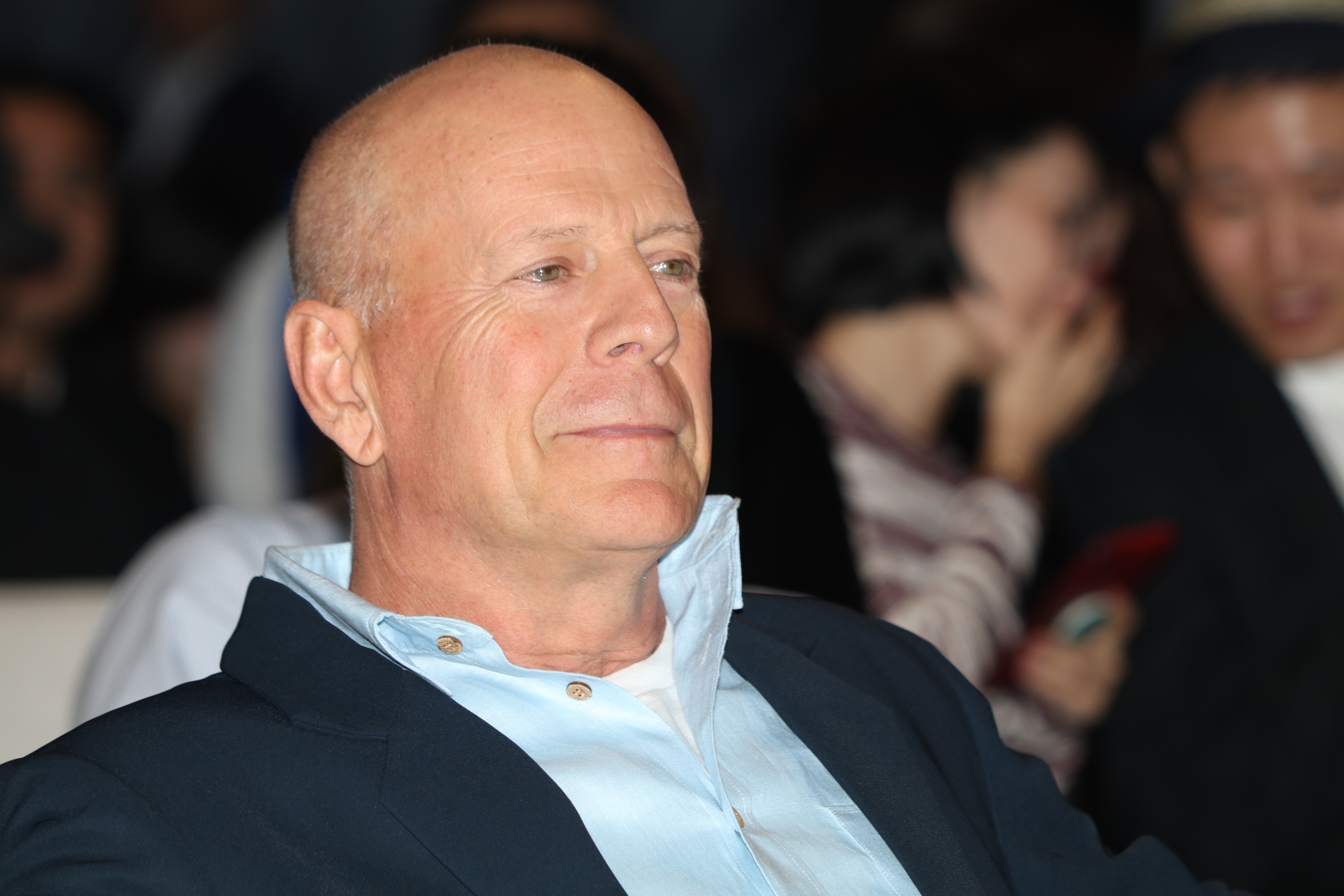 Bruce Willis smiles slightly, wearing a light shirt under a dark blazer. Blurred individuals in the background are engaged in conversation