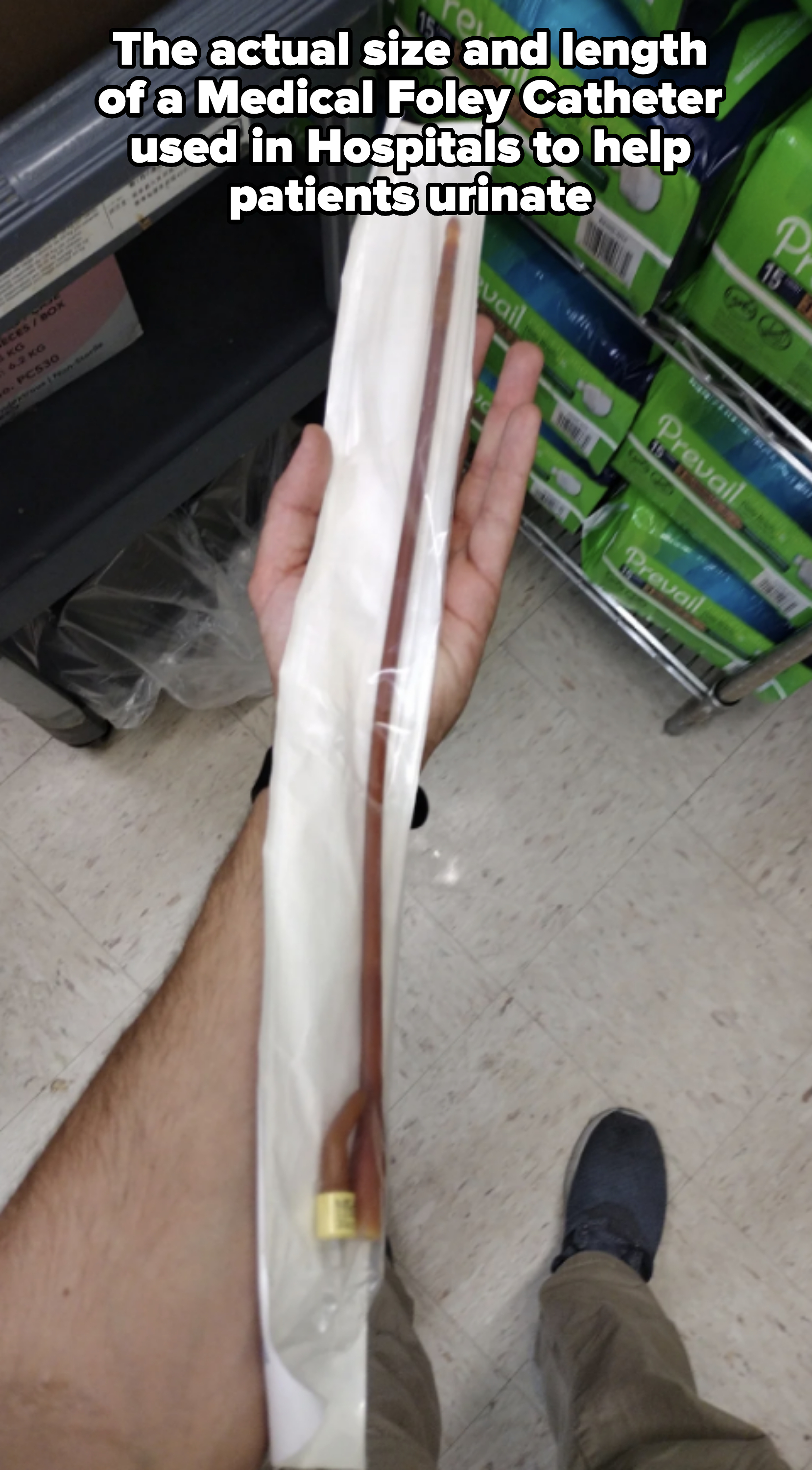 A hand holds a sealed, long, thin medical catheter near shelves stocked with incontinence products