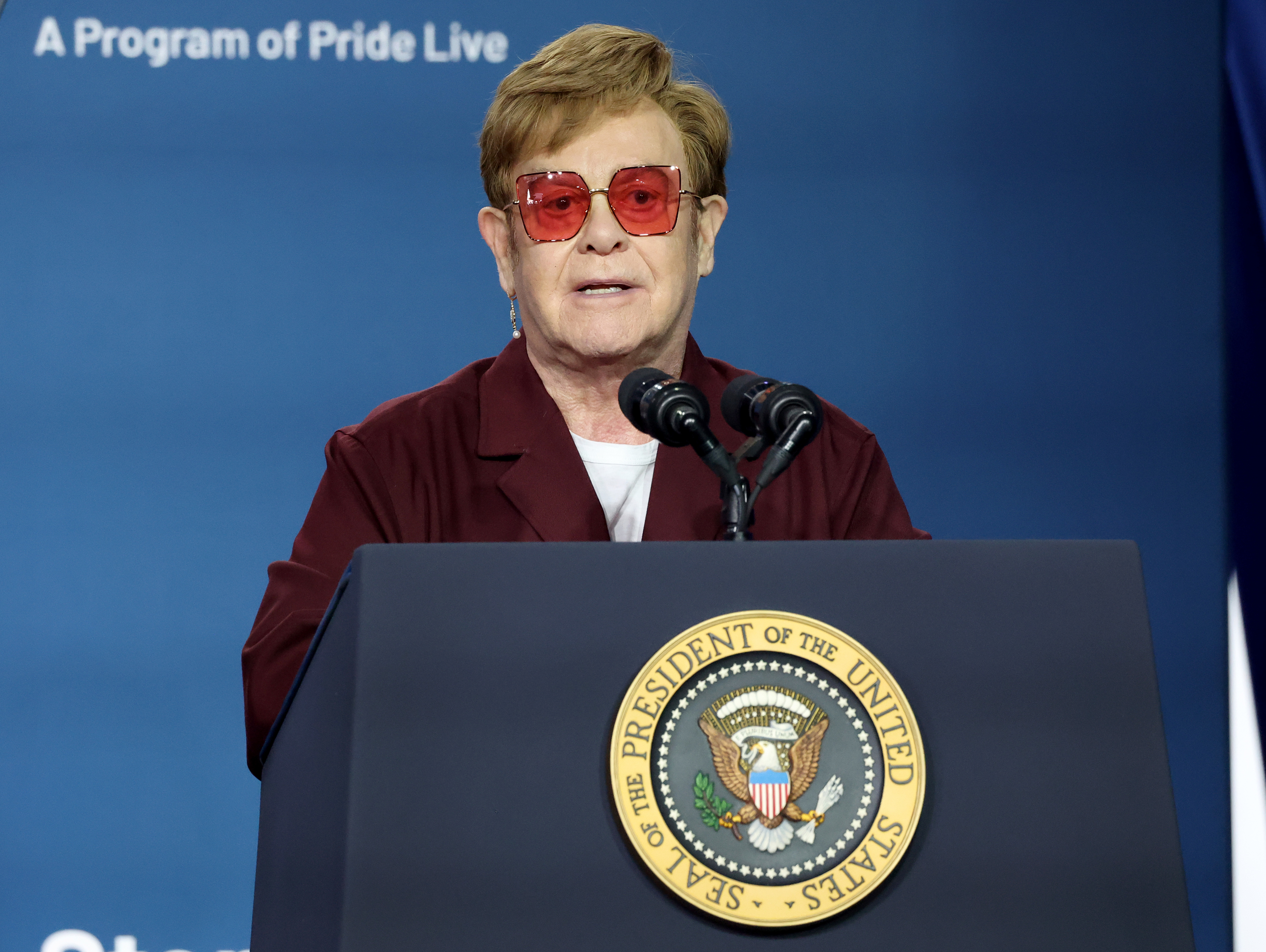 Elton John speaks at a podium with the emblem of the President of the United States, wearing red-tinted glasses and a dark suit