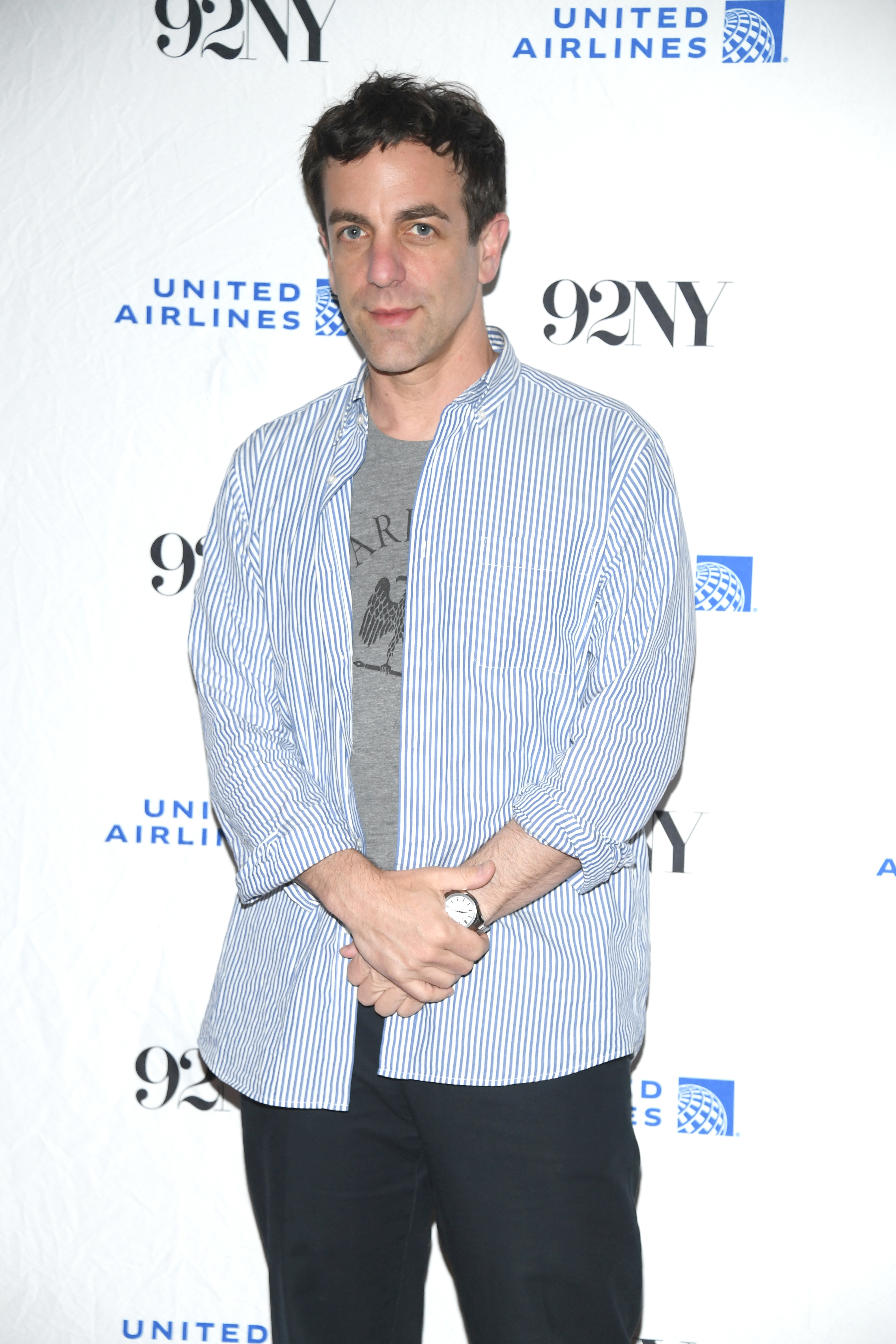 B.J. Novak stands in front of a backdrop with logos for United Airlines and 92NY, wearing a striped shirt over a graphic T-shirt