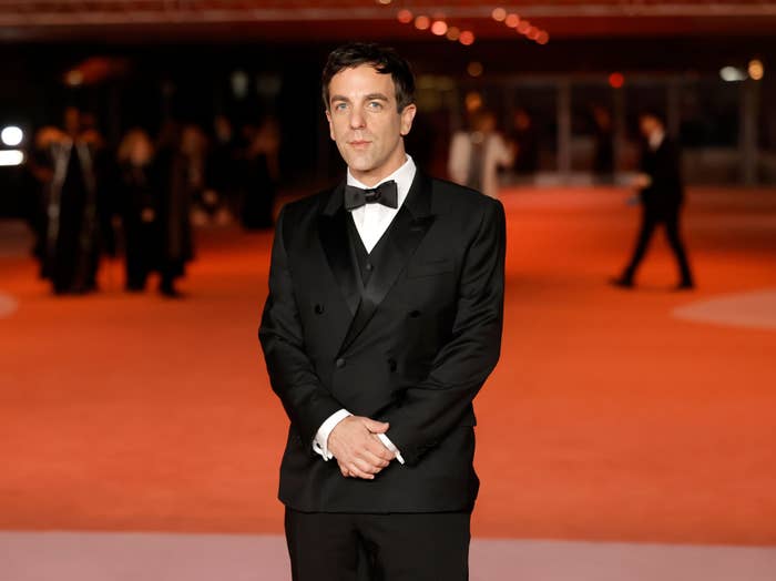 B.J. Novak stands on a red carpet wearing a black tuxedo