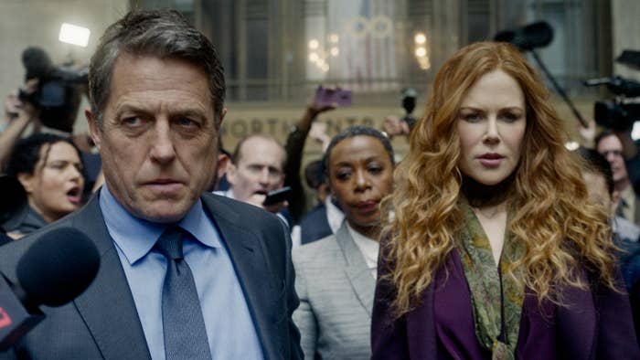 Hugh Grant in a suit and Nicole Kidman in a stylish outfit stand amidst a crowd of reporters and photographers, looking serious