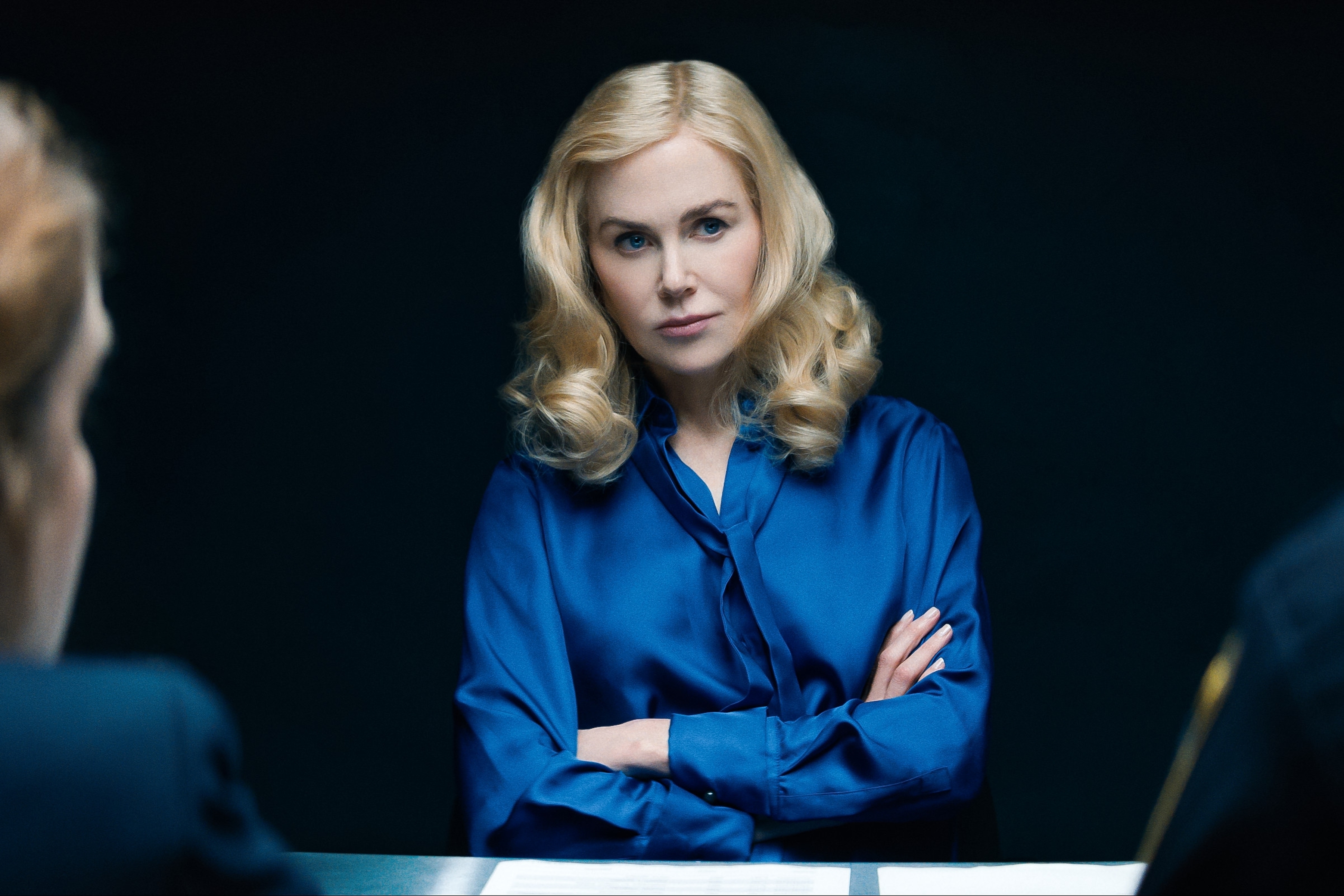 Nicole Kidman seated with arms crossed, wearing a satin blouse, facing two people at a table in a dimly lit room