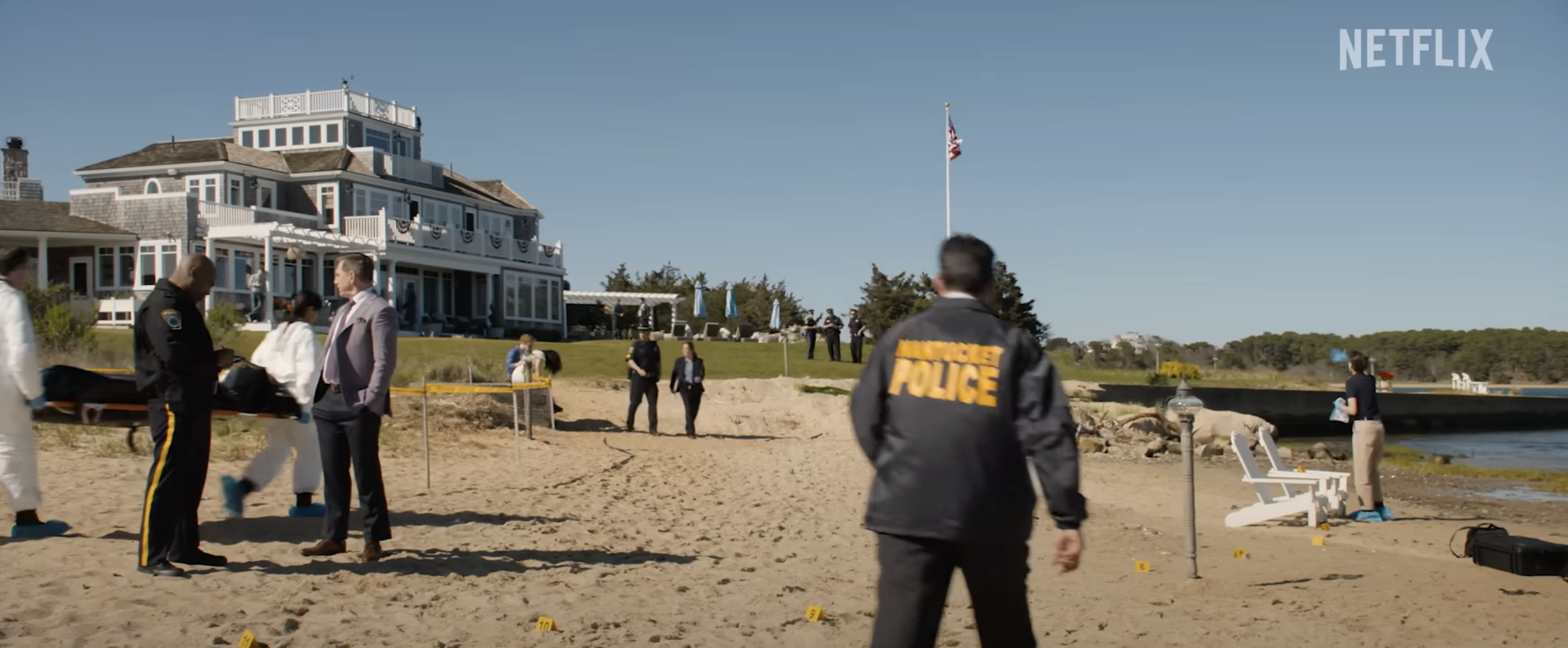 A crime scene near a beach, with investigators and police officers. A large house is in the background. Text in the top right reads &quot;NETFLIX.&quot;