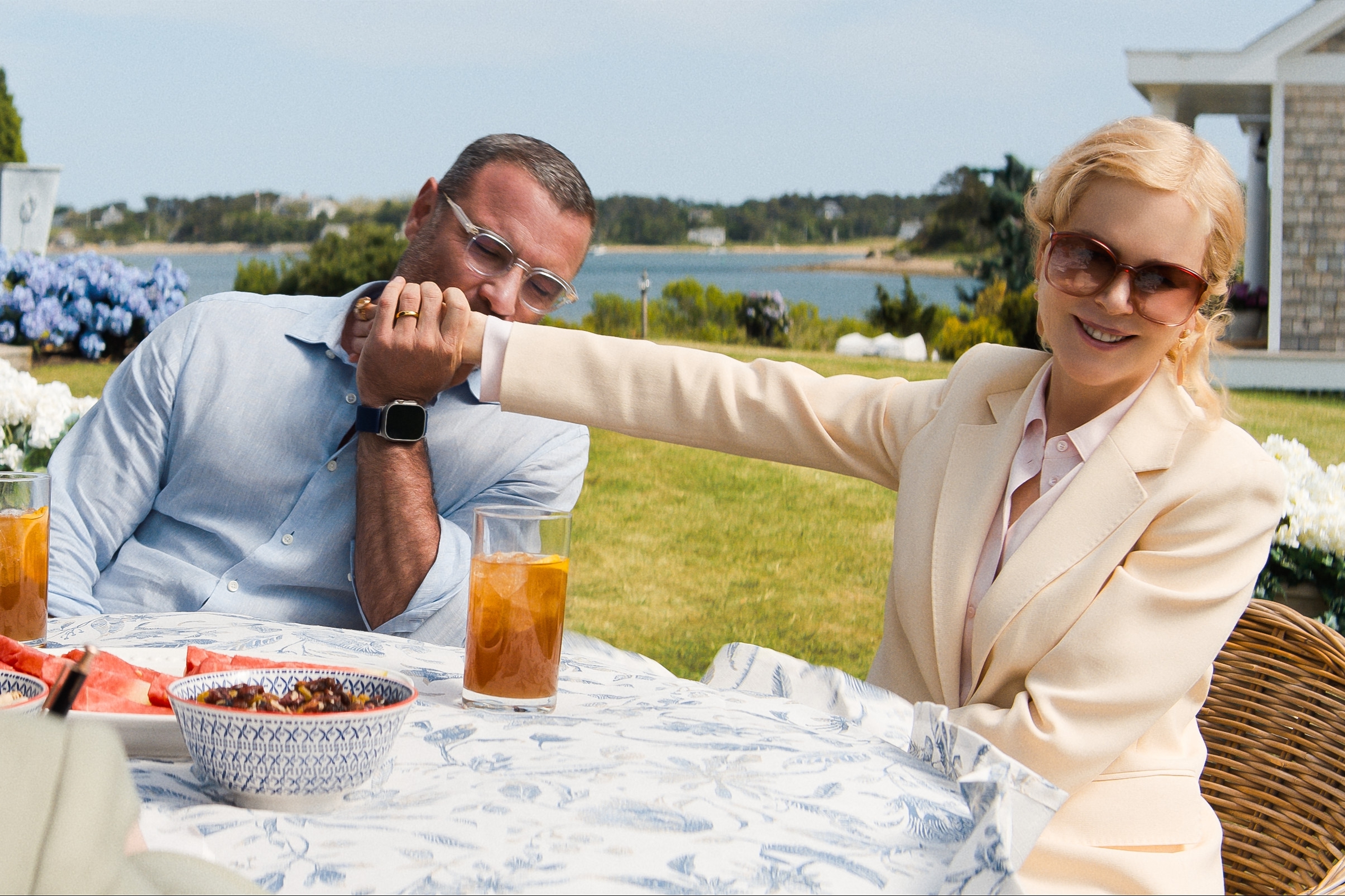 Liev Schreiber and Nicole Kidman are seated outdoors at a table. Liev is wearing a blue shirt and a smartwatch, while Nicole wears sunglasses and a light-colored blazer