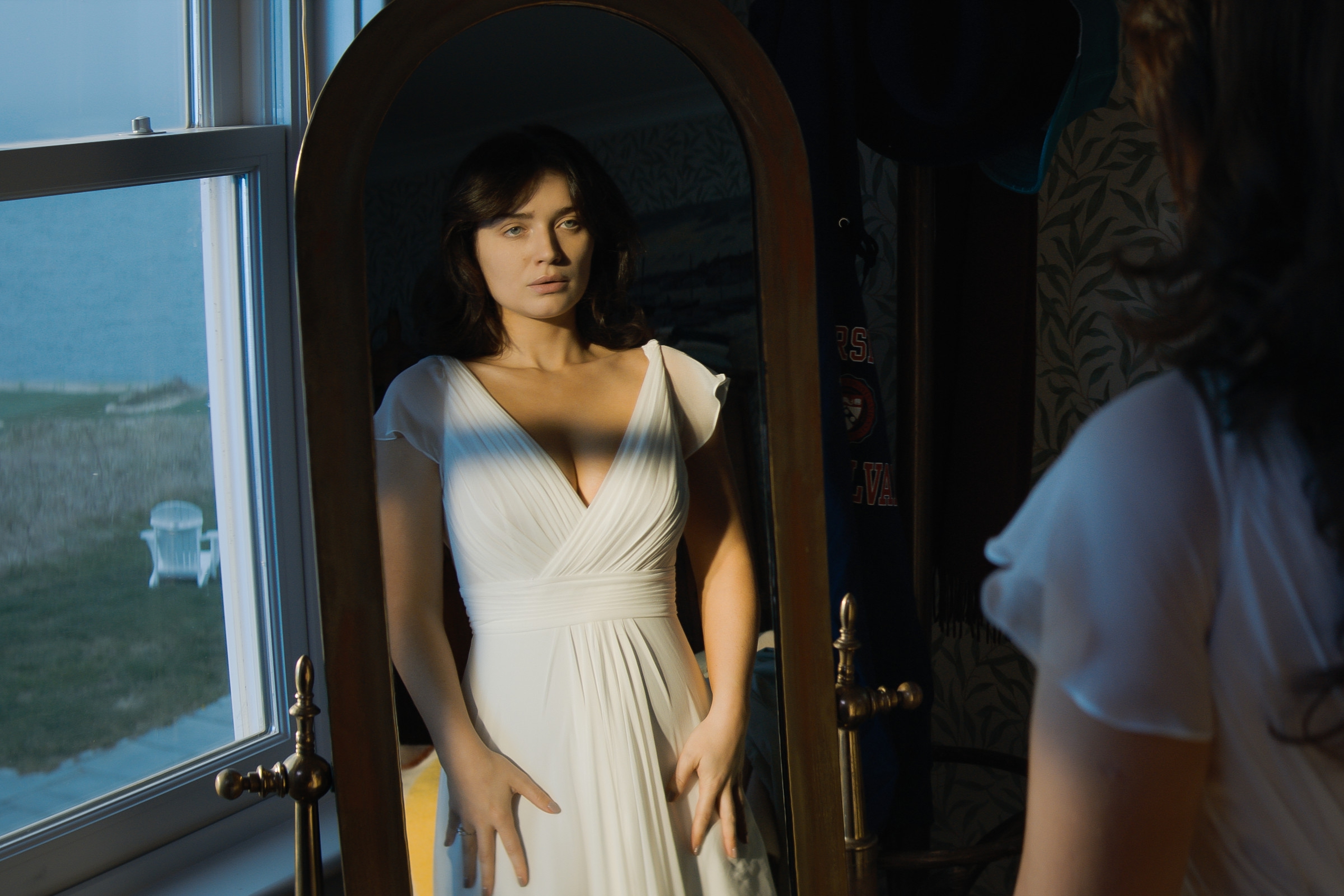 Eva in a dress looks into a tall mirror, standing by a window with a view of an outdoor scene