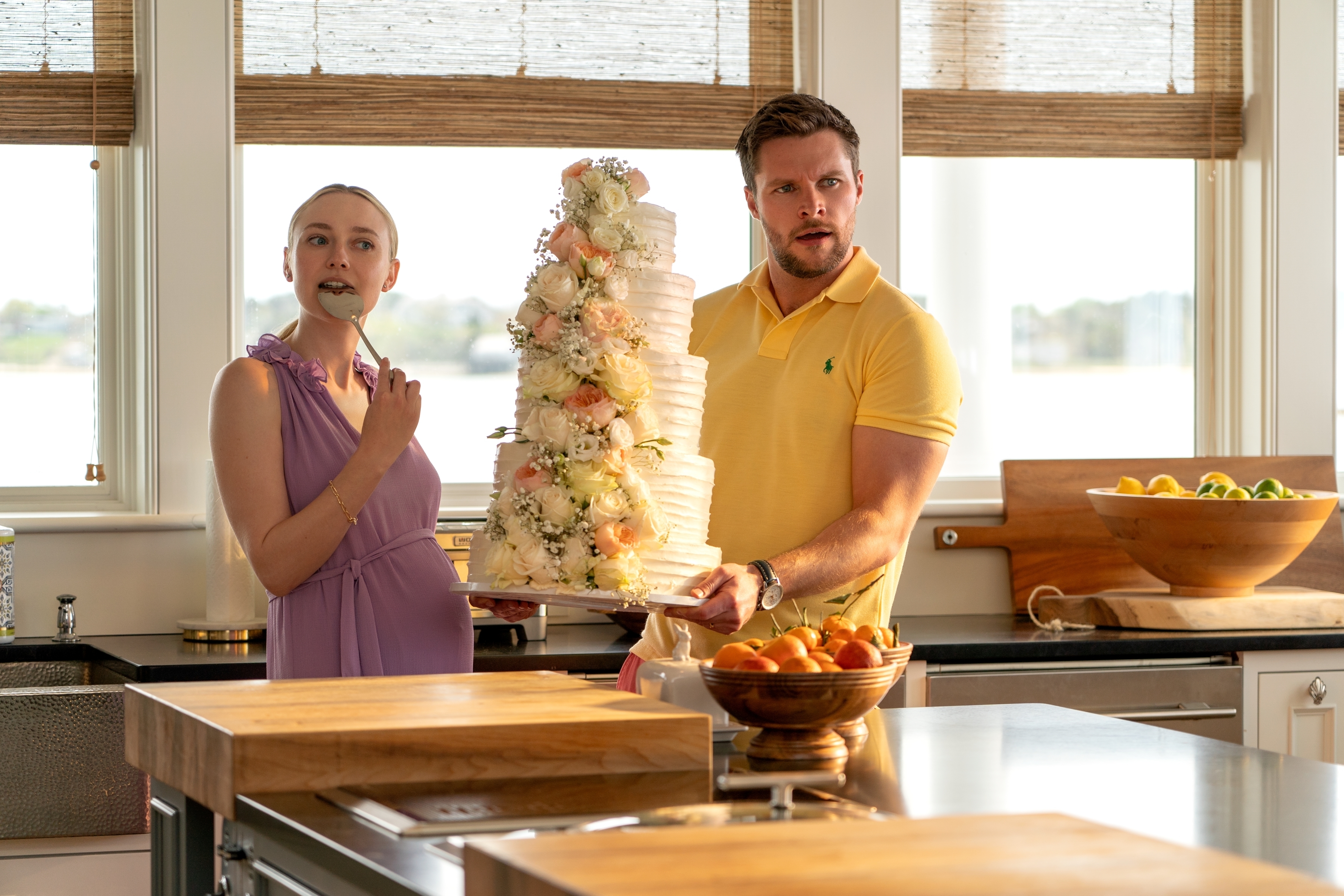 Dakota Fanning wearing a sleeveless dress and Jack Reynor in a polo shirt, holding a tall wedding cake in a kitchen