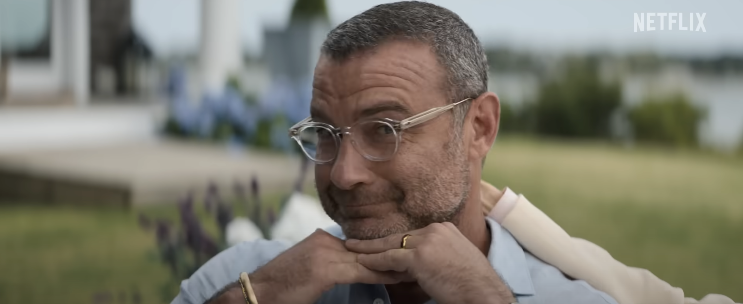 Liev Schreiber smiles mischievously with hands under his chin while wearing glasses and a light shirt