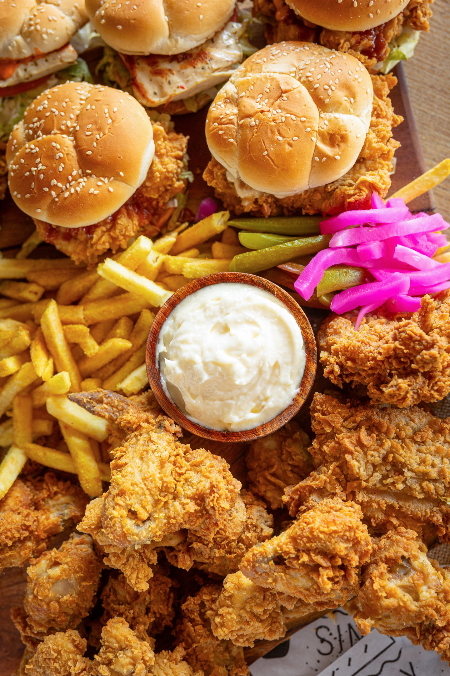 An assortment of fried chicken, sandwiches, fries, and pickled vegetables on a platter with a bowl of dipping sauce in the center