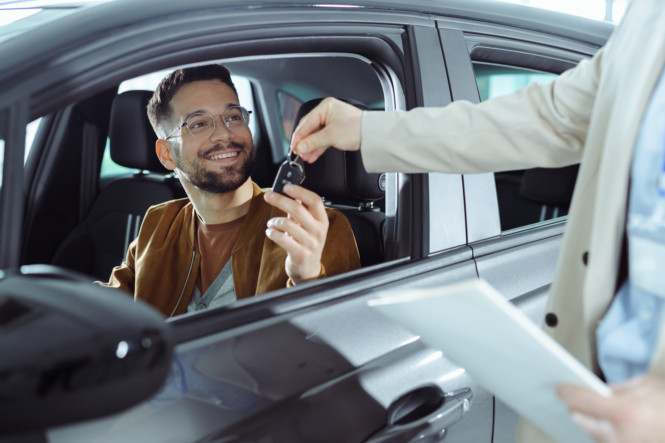 A man in a car receives keys from the hand of a person standing outside the vehicle. Both appear content