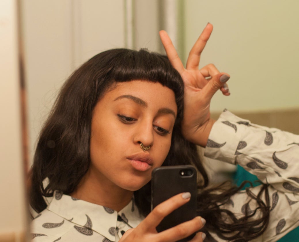 Person takes a selfie in a bathroom mirror, making a peace sign with one hand. They have long, dark hair and wear a patterned sweatshirt with a small, distinct print