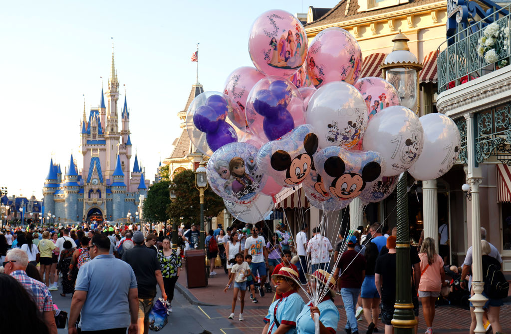 A bustling Disney park with guests walking near Cinderella Castle and a balloon vendor holding various character balloons, including Mickey Mouse and Frozen characters