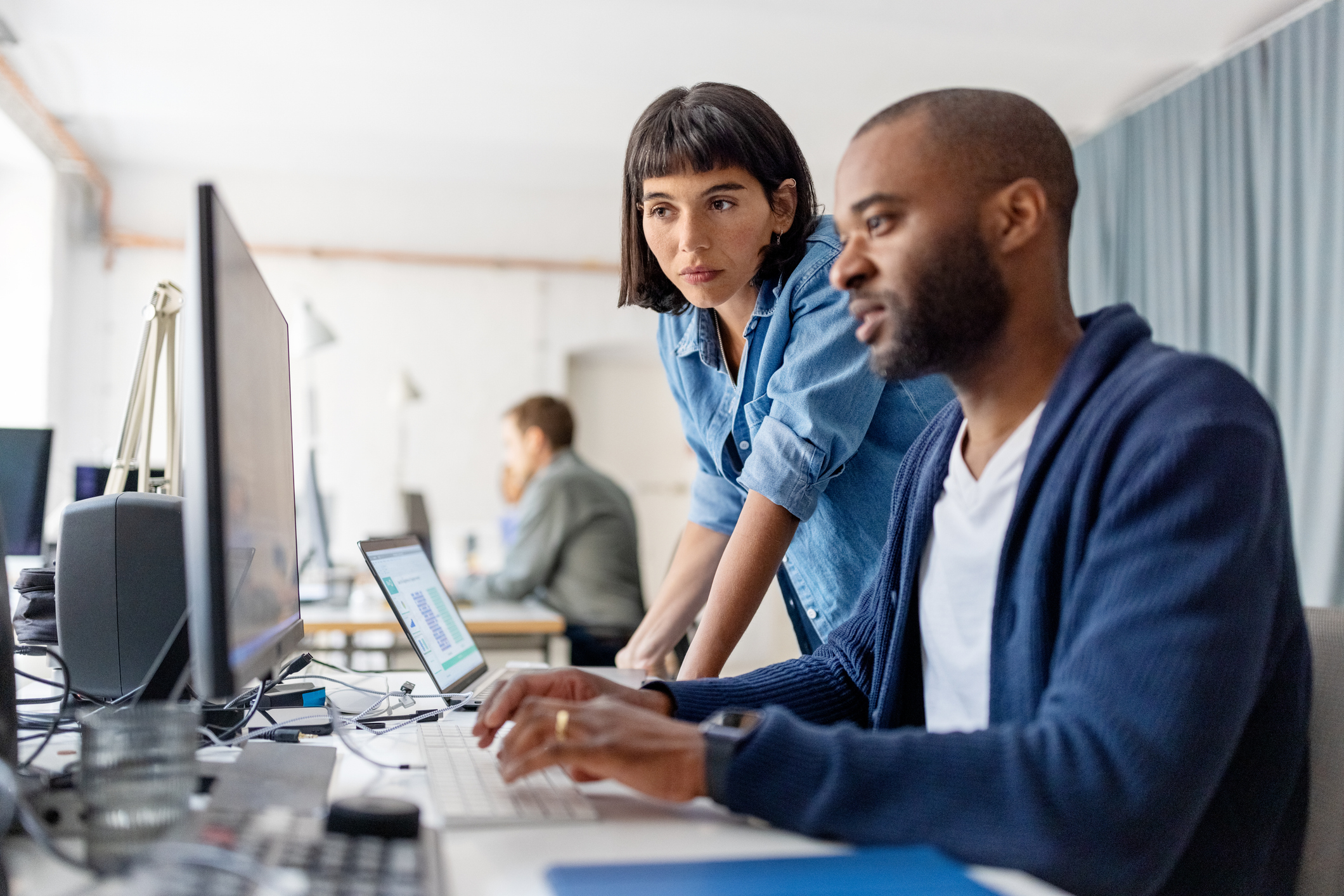 A woman and a man are working at a desk with computers. The woman is standing and looking at the man&#x27;s screen. Another person is in the background