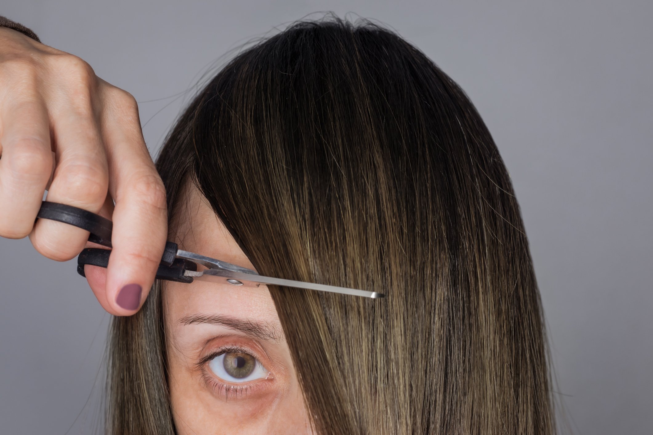 A close-up of a person cutting their own bangs with scissors. The person&#x27;s eye and hand holding the scissors are visible