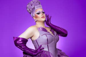 Drag queen in a glamorous, structured outfit and long gloves poses confidently, with elaborate makeup and styled hair against a purple background