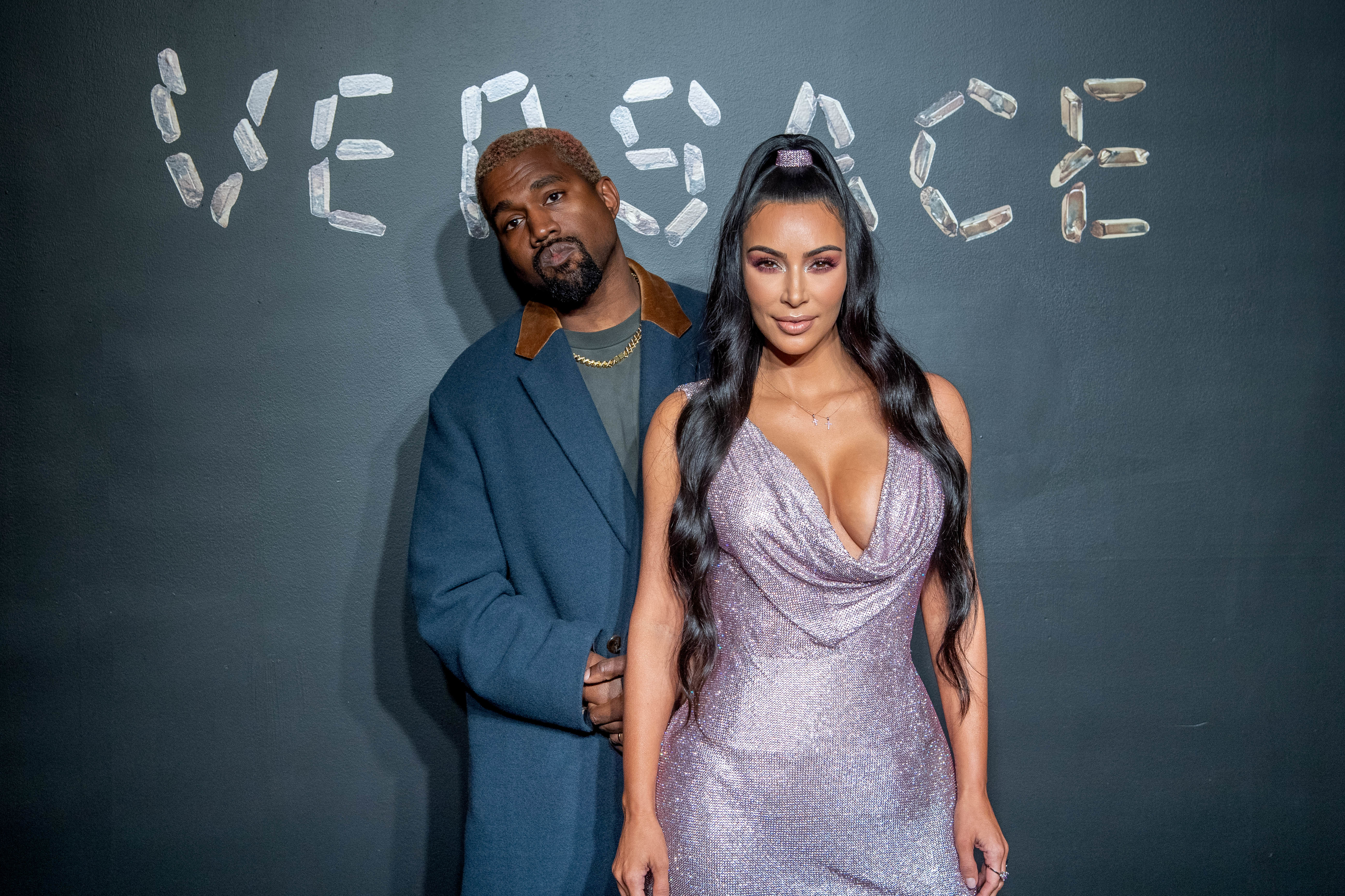 Kanye West in a gray coat and Kim Kardashian in a shimmering dress pose together at a Versace event. Versace text in the background