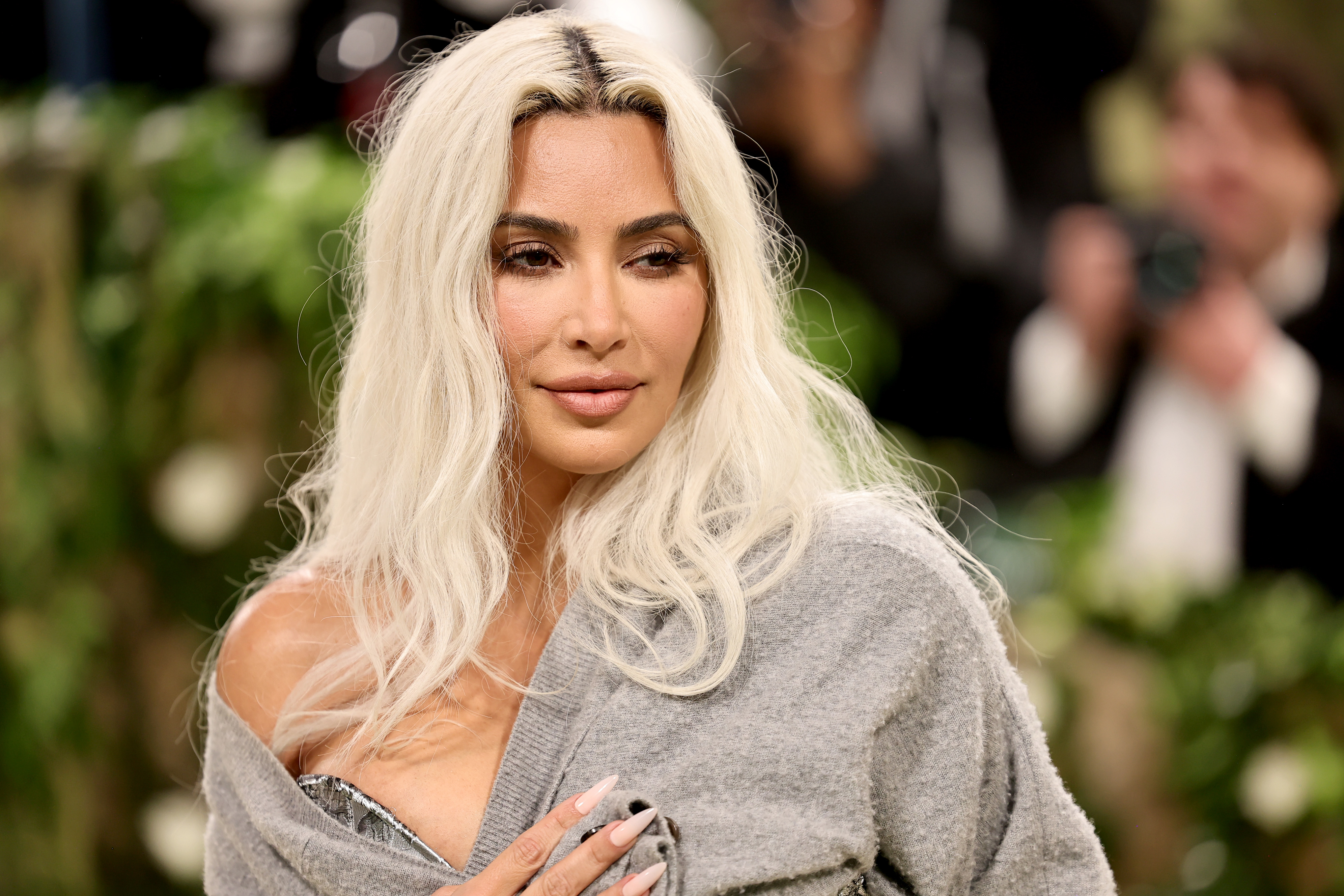 Kim Kardashian poses outdoors wearing a gray shawl, looking to the left, with soft makeup and wavy platinum blonde hair cascading over her shoulders