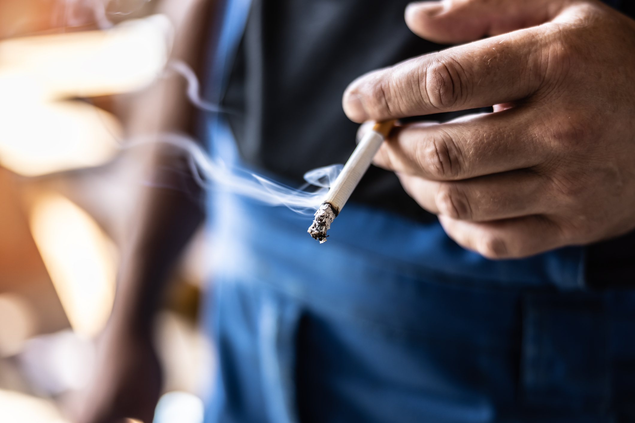 A person holding a lit cigarette with smoke rising in the air. The person&#x27;s face and body are not visible