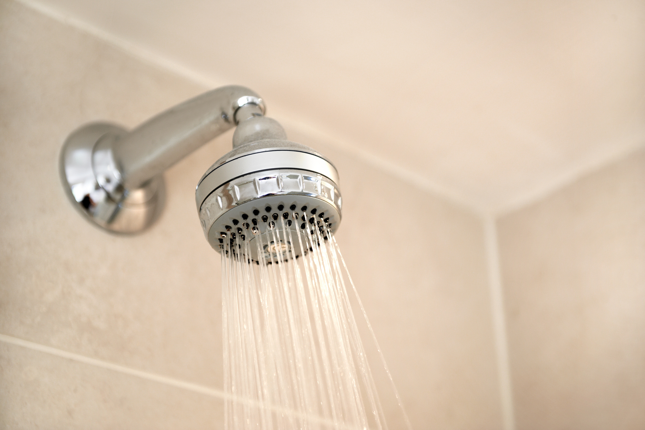 A close-up of a showerhead with water spraying out, mounted on a beige tiled wall