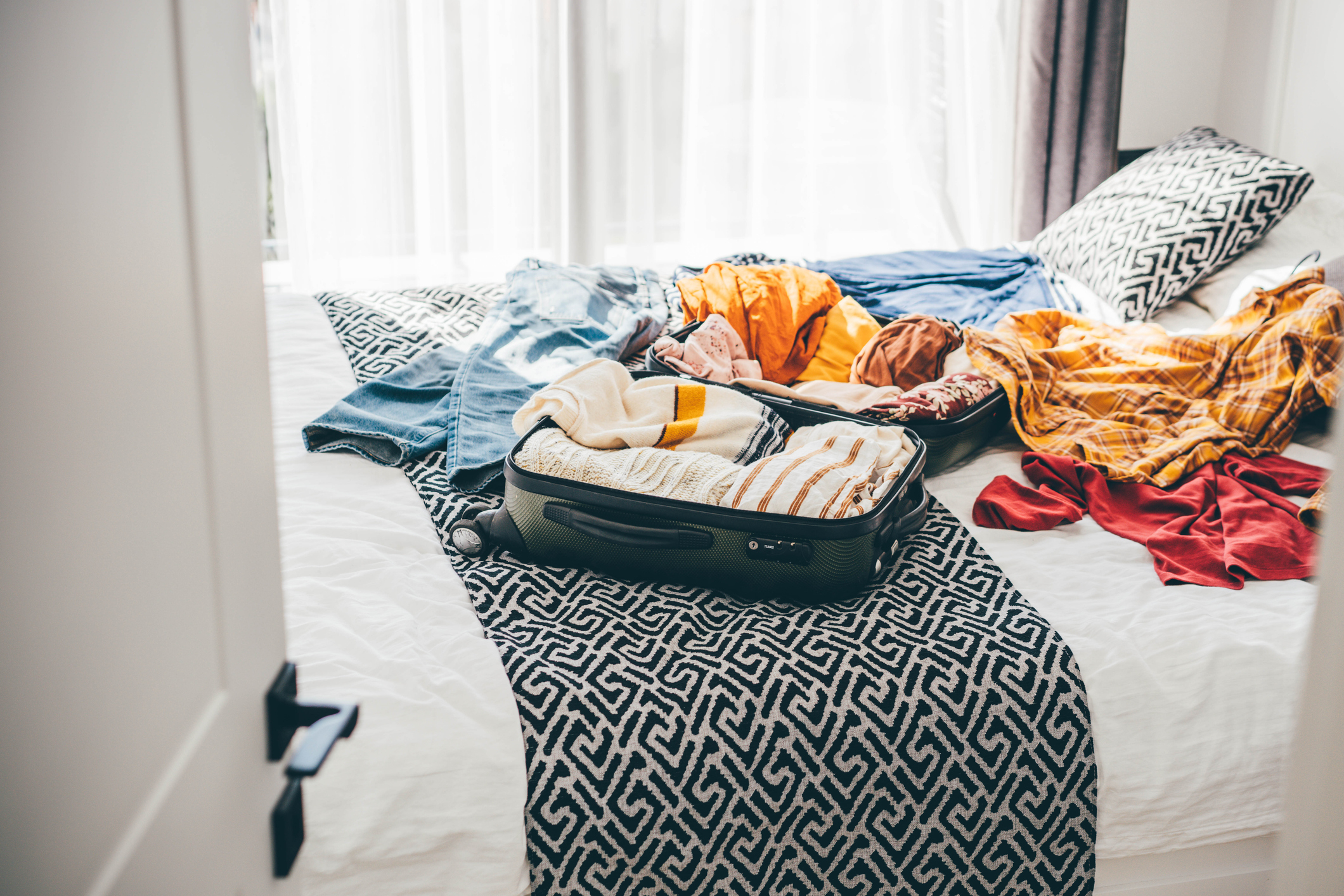 An open suitcase with clothes spilling out is placed on a bed in a well-lit room with patterned bedding