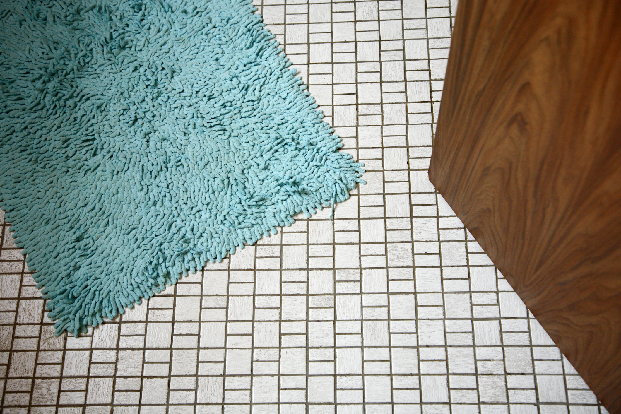 Close-up of a corner in a bathroom with a fluffy bath mat on tiled flooring next to a wooden cabinet