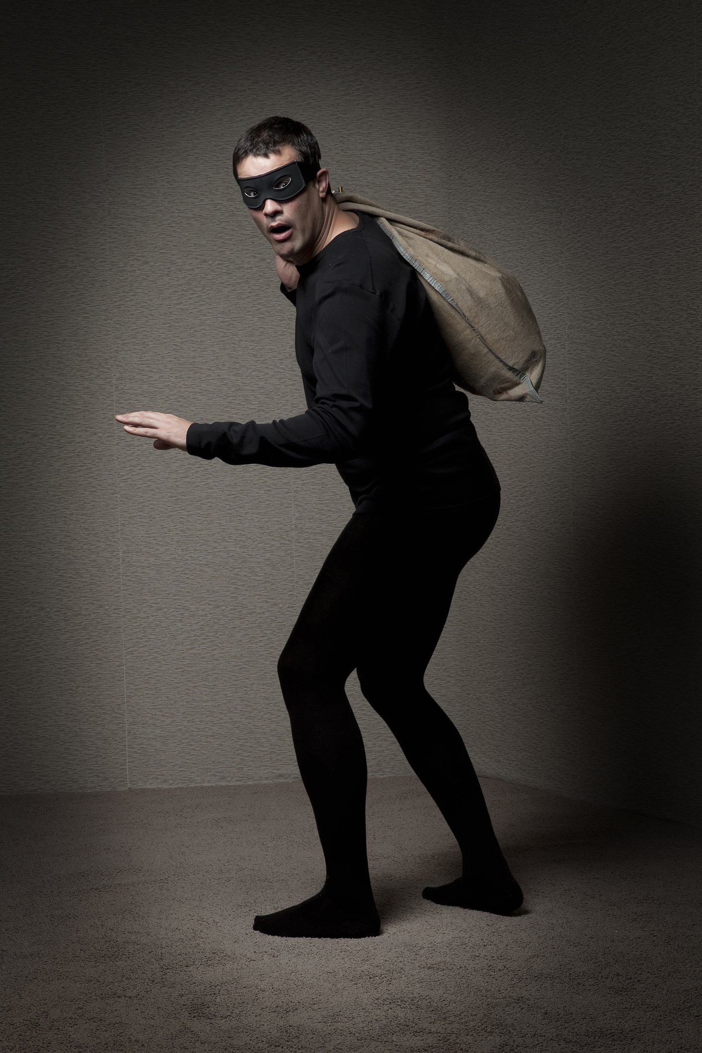 Man dressed as a burglar in black clothes and mask, carrying a sack over his shoulder, striking a sneaky pose in a sparse room