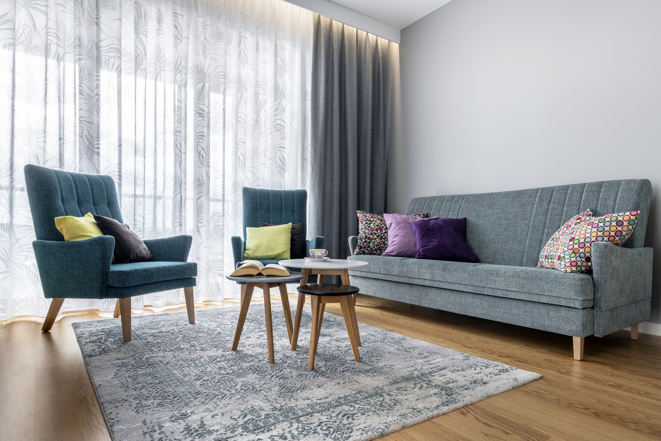 A cozy living room with two blue armchairs, a blue sofa, colorful pillows, a small coffee table, an open book, and a cup next to it, on a patterned rug