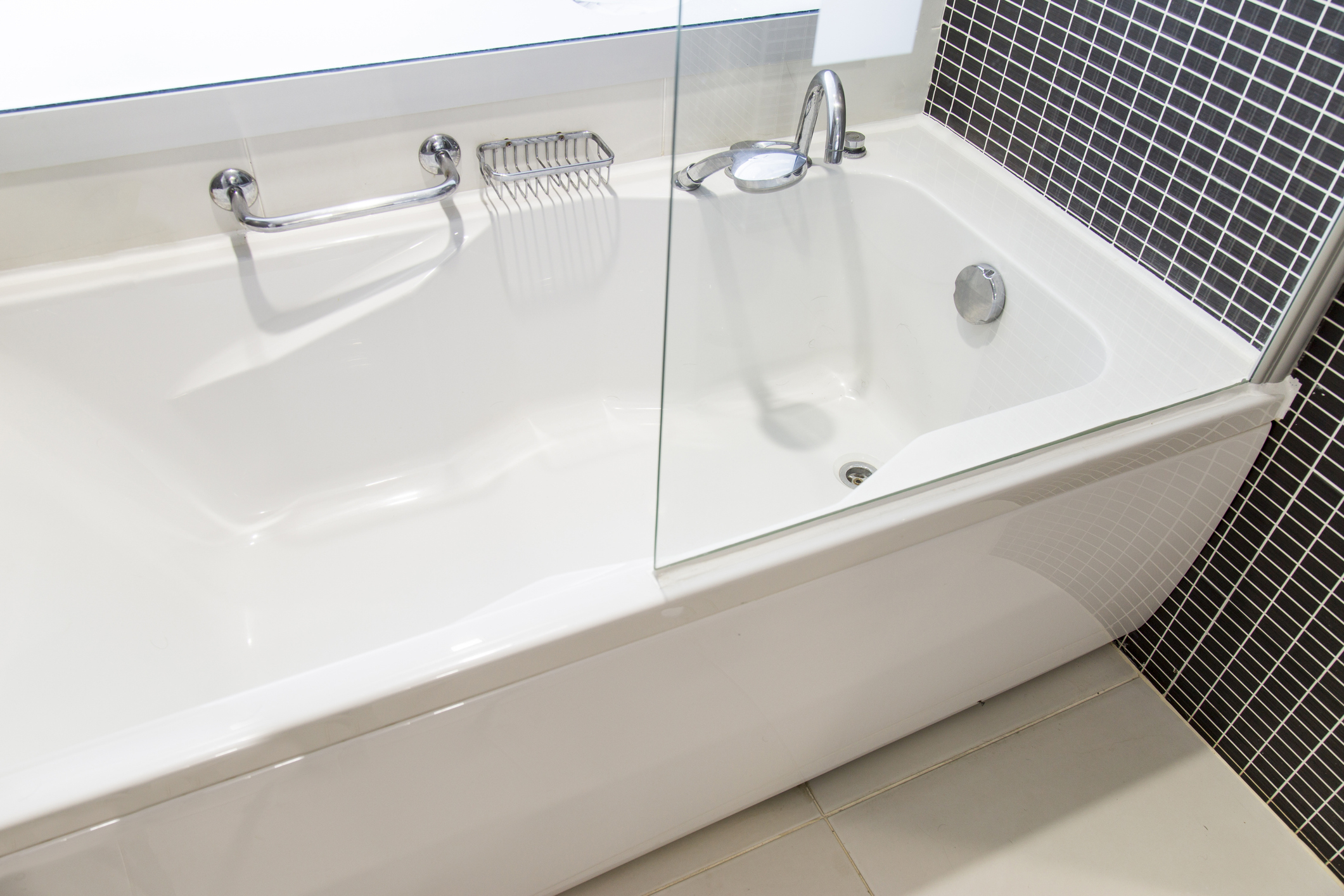 White bathtub with a glass shower door, metal faucet, and a black tiled wall on the right side. A metal grab bar and soap holder are mounted on the tub wall