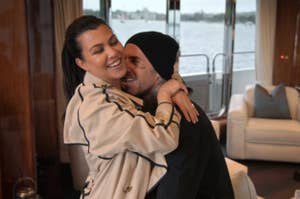 Kourtney Kardashian and Travis Barker are embracing and smiling. Kourtney is wearing a trench coat, and they are indoors with a window view in the background