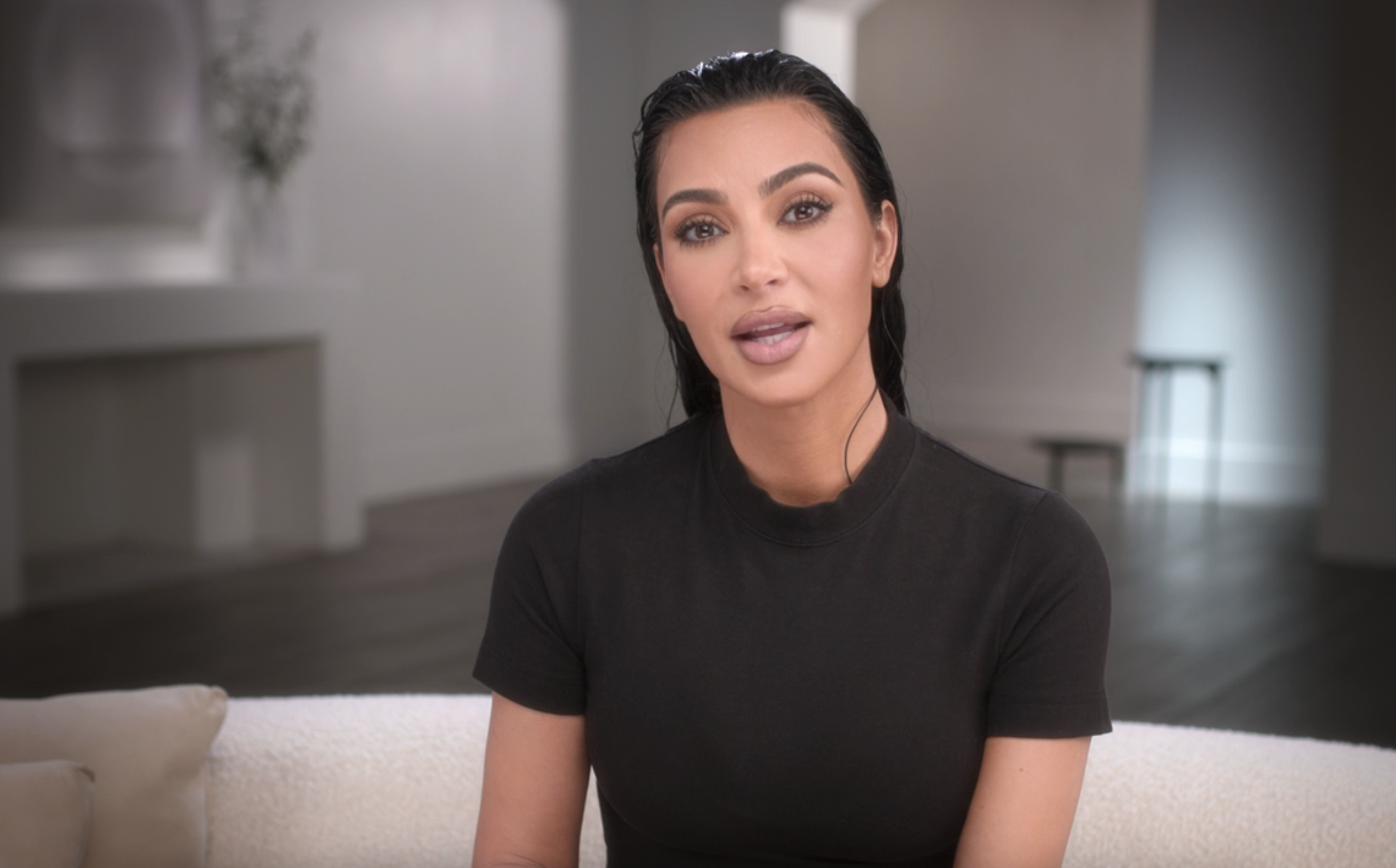 Kim Kardashian sitting on a couch, wearing a simple black top, with a minimalist background behind her