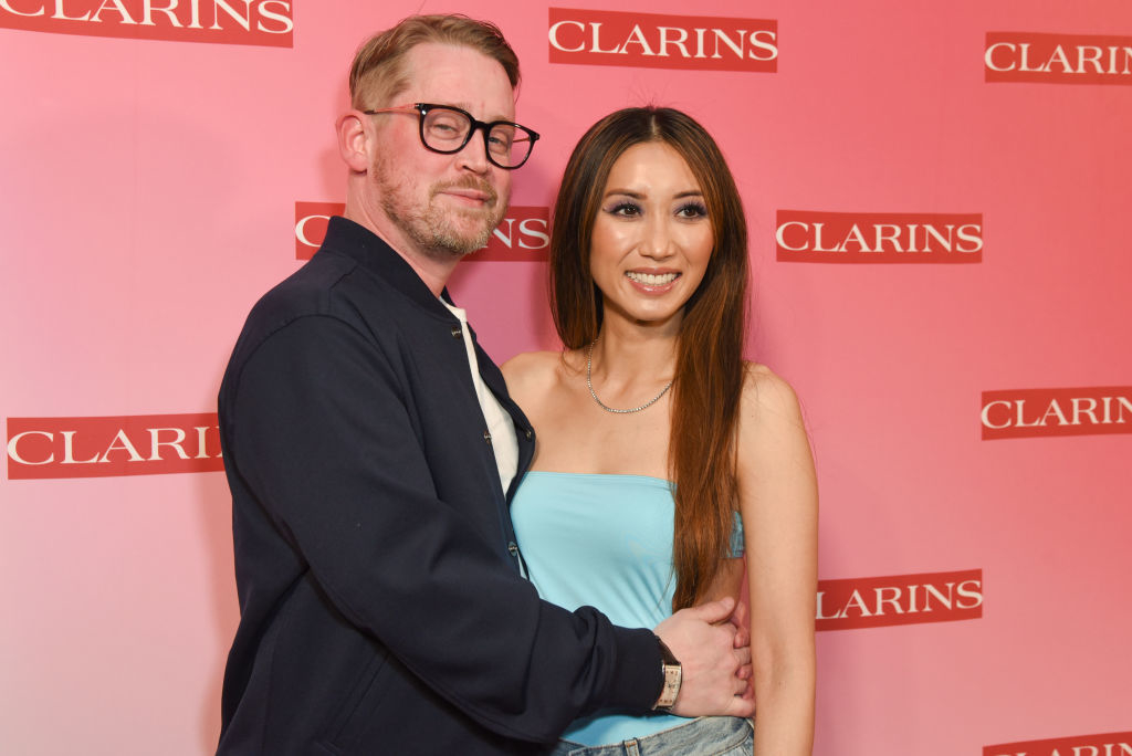 Macaulay Culkin and Brenda Song pose together at a CLARINS event. Macaulay wears glasses, a dark jacket, and a white shirt; Brenda wears a strapless top