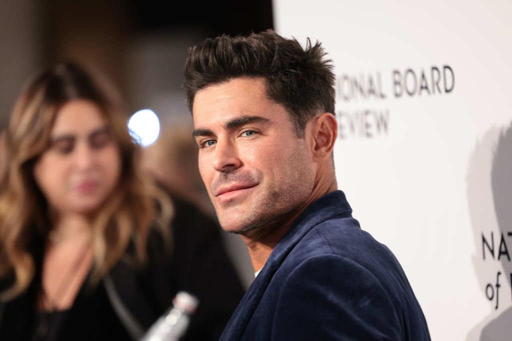 Zac Efron, in a stylish suit jacket, poses for the camera at a National Board of Review event. A person is blurred in the background