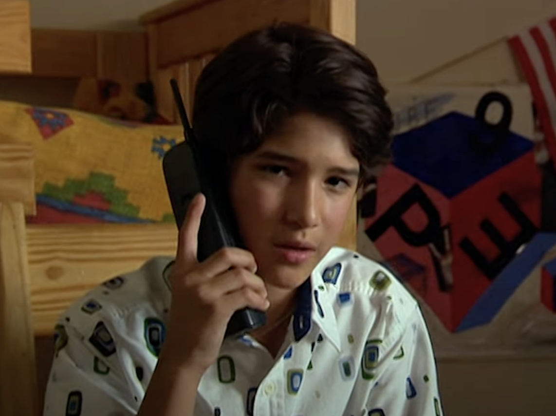 Young boy holding a large cell phone, wearing a patterned shirt, sitting in a bedroom