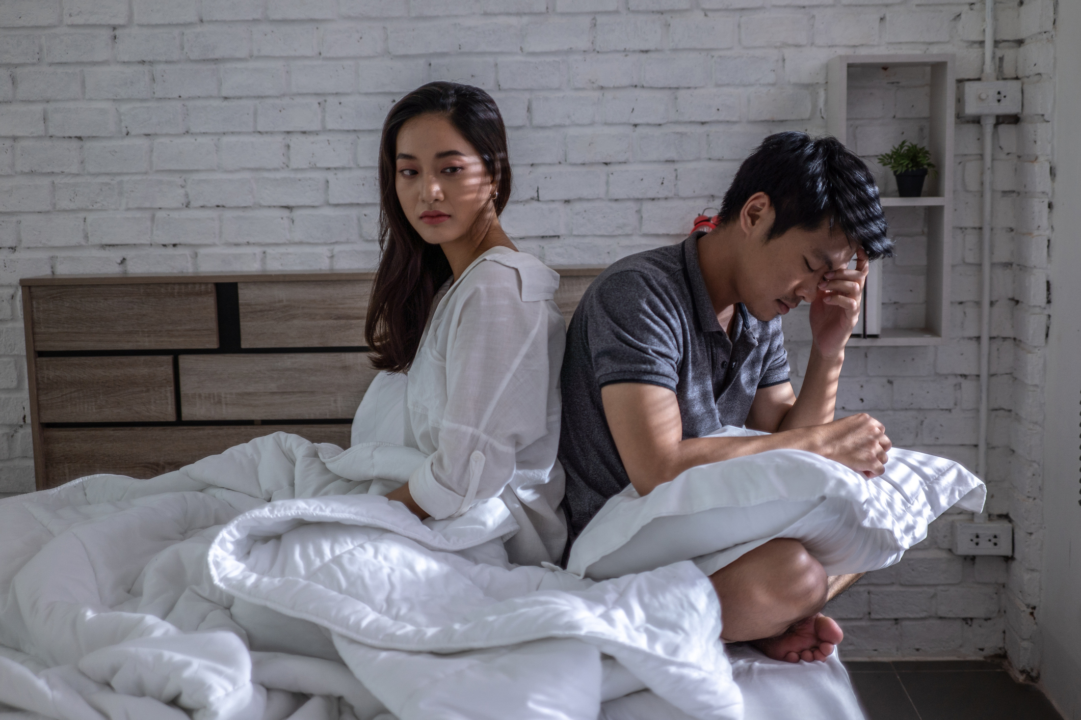 A woman and a man sit on a bed with white bedding. The woman looks away, appearing thoughtful, while the man holds his head with one hand, looking distressed