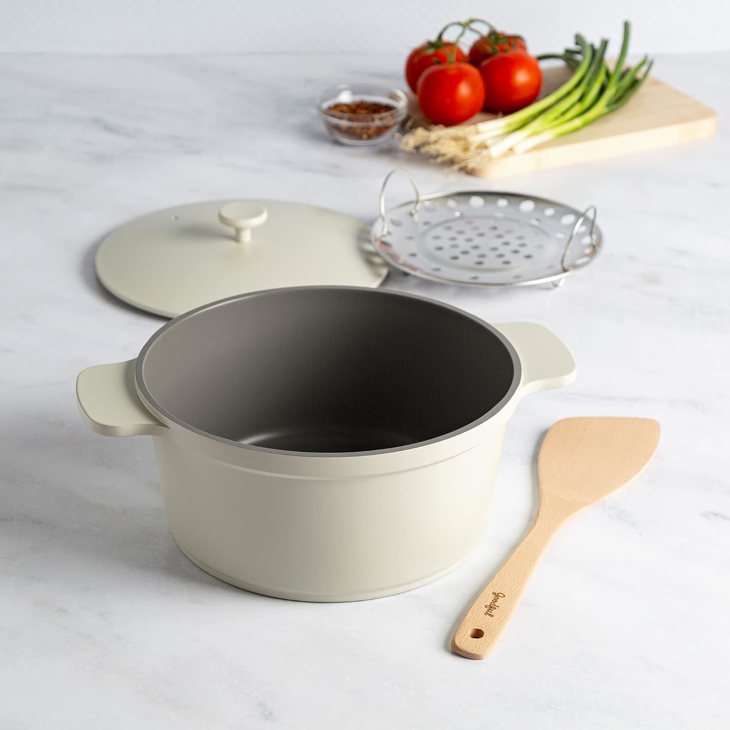 Large cooking pot with lid removed, wooden spatula, steamer insert, tomatoes, green onions, and dried spices on a cutting board in the background