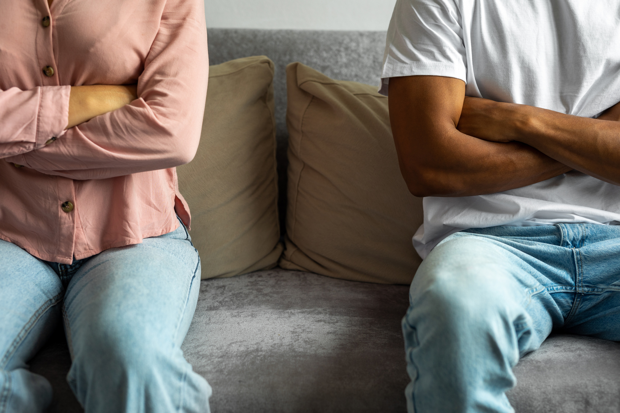 Two people sit on a couch with their arms crossed, facing slightly away from each other, suggesting a disagreement or conflict