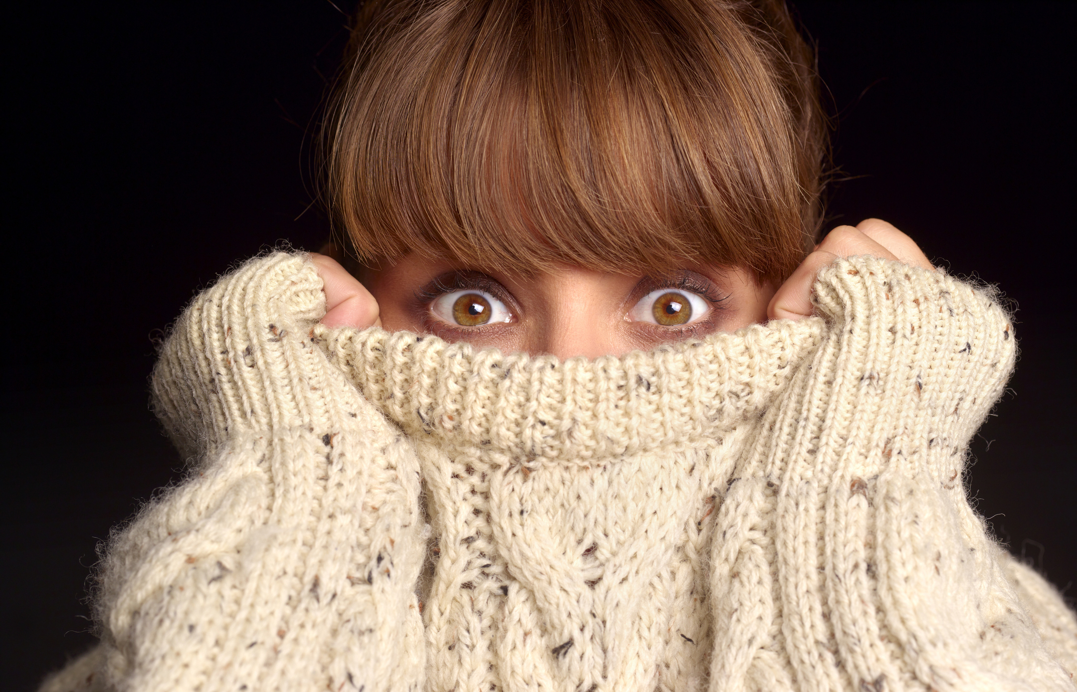 A woman partially covers her face with a cozy, knit sweater, showing only her wide eyes and bangs