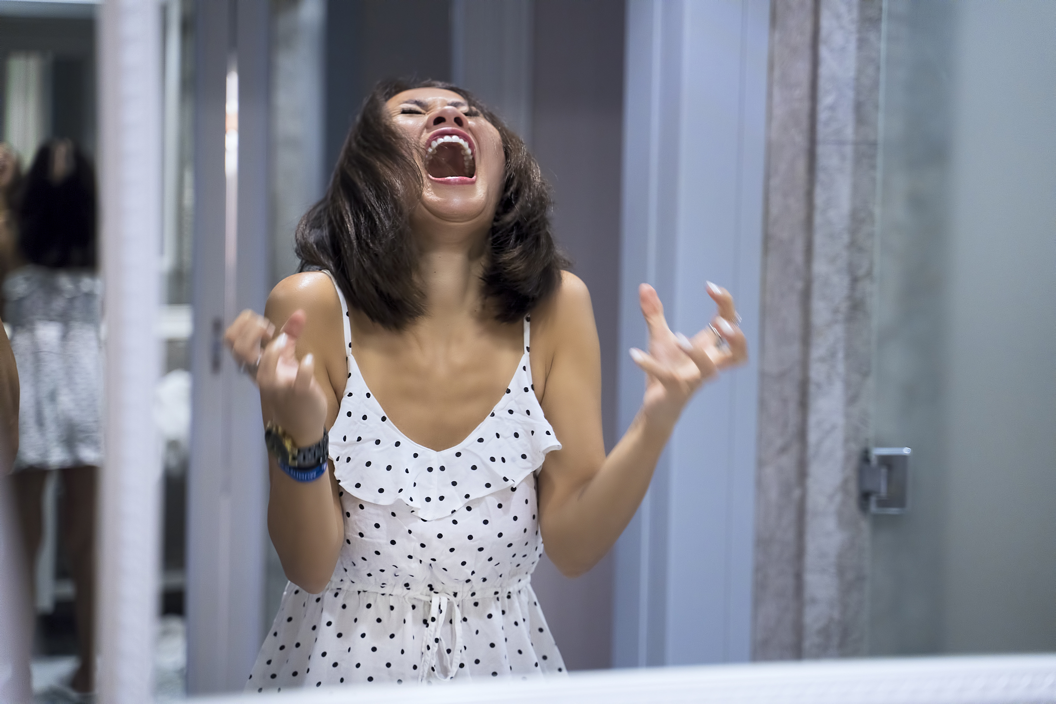 A woman with shoulder-length hair, wearing a white dress with black polka dots, is dramatically laughing or yelling in front of a mirror