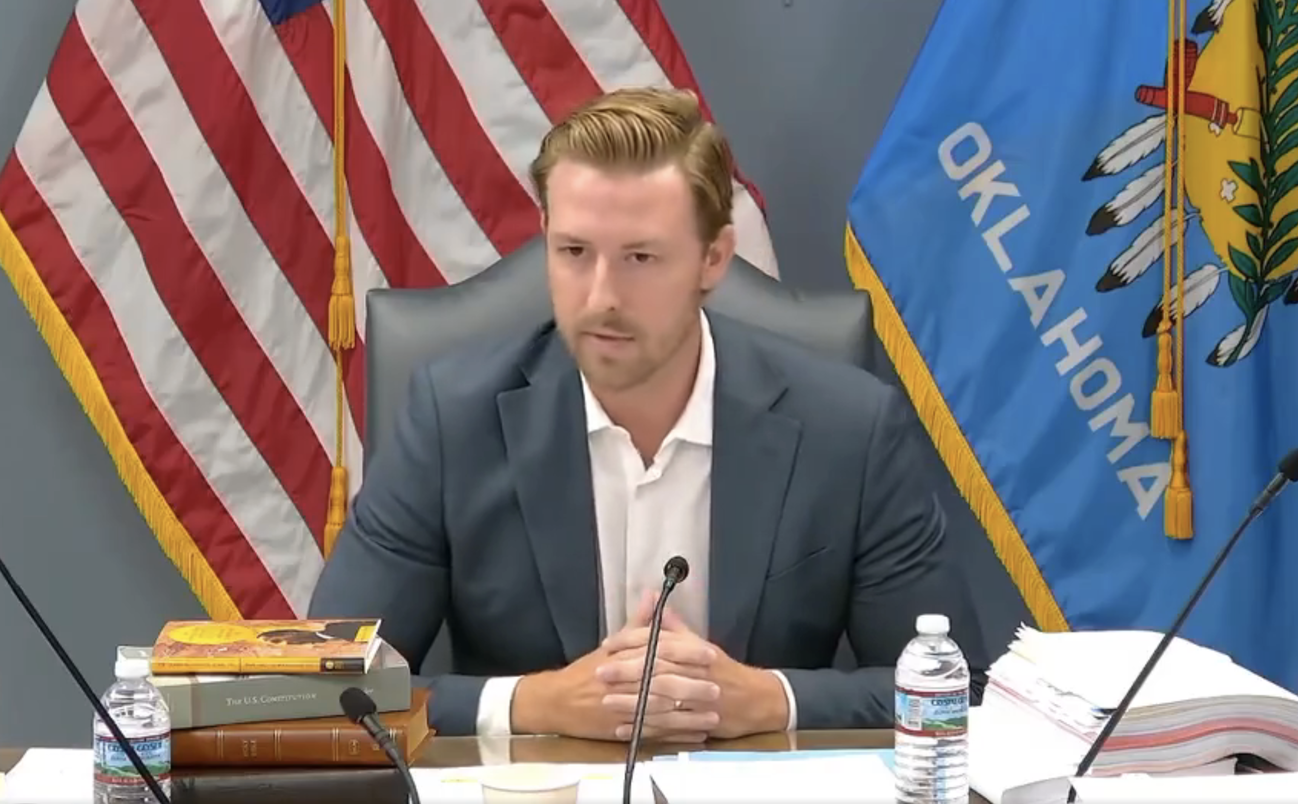 A man sits at a desk with books, a water bottle, and documents in front of him, speaking at a meeting. Two American flags and an Oklahoma flag are behind him