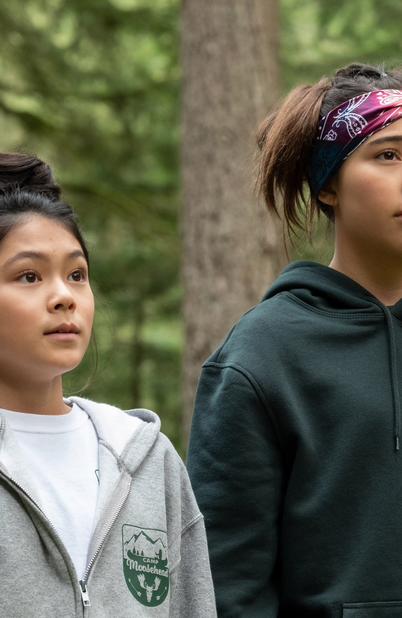 Dawn wearing a hoodie with a forest emblem, standing in a wooded area