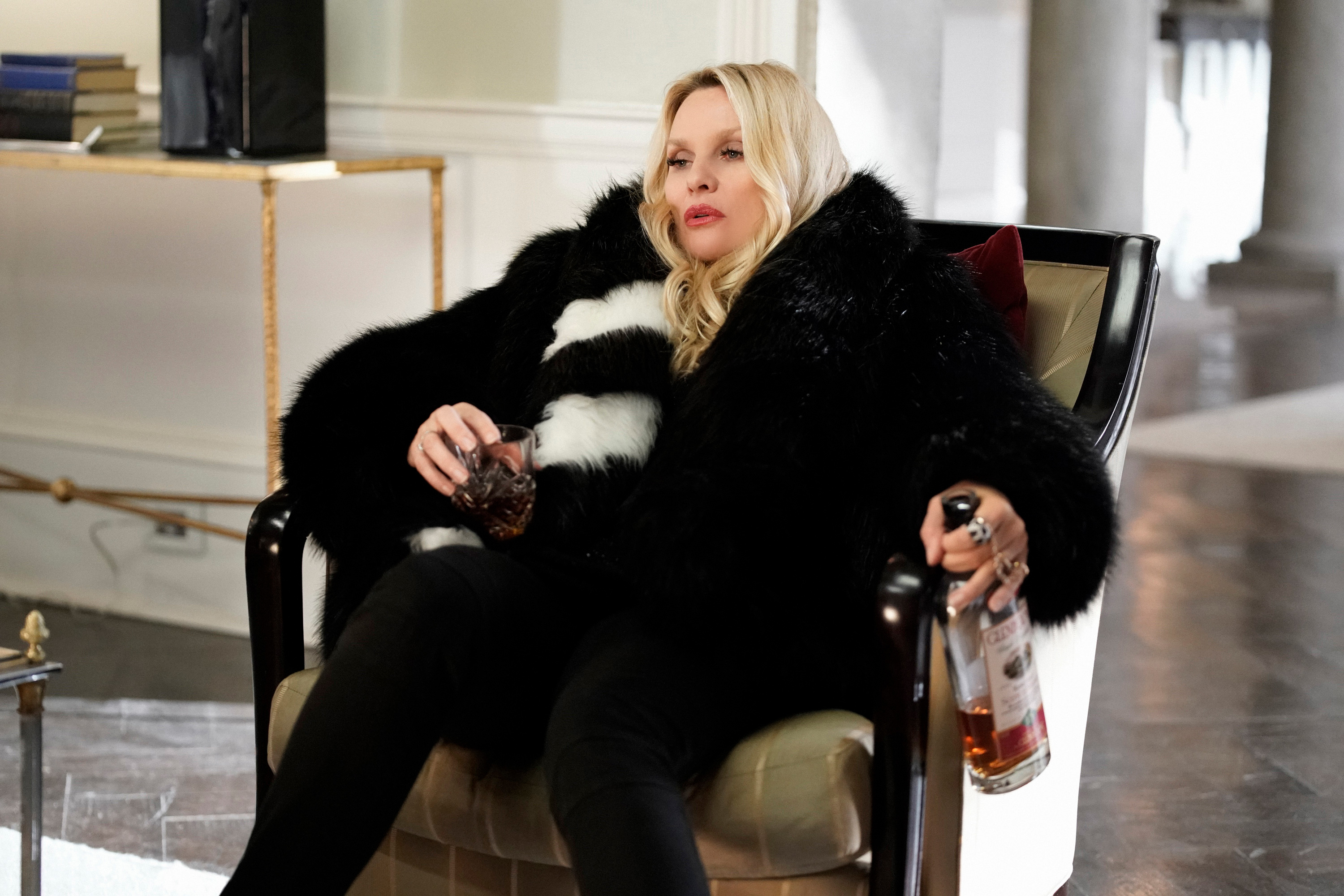 Alexis in a plush chair holding a bottle of liquor in one hand and a glass in the other, dressed in a luxurious fur coat, appearing relaxed