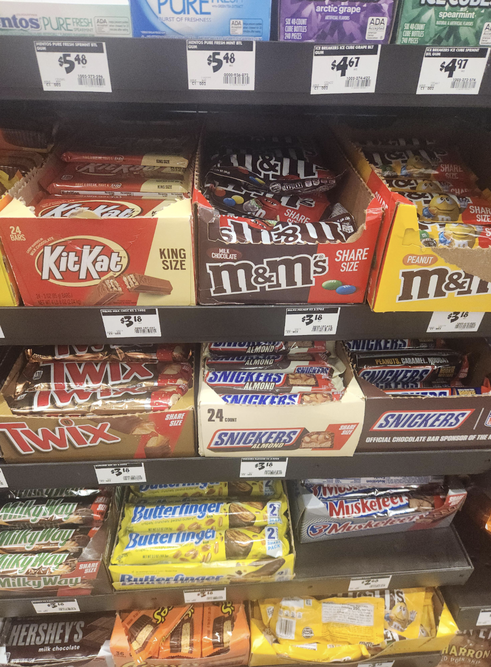 Store shelf filled with various candy bars including KitKat, M&amp;amp;M&#x27;s, Twix, Snickers, Butterfinger, 3 Musketeers, Hershey&#x27;s, and others